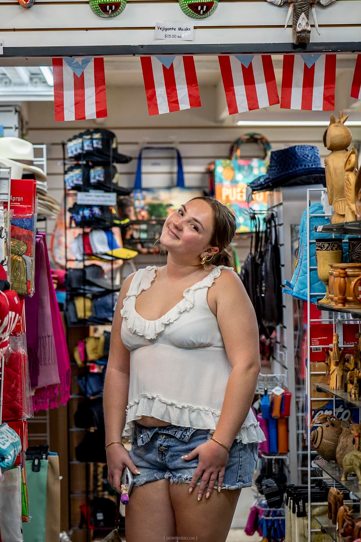 Young woman in white crop top and denim shorts smiling in a colorful souvenir shop with Puerto Rican flags.