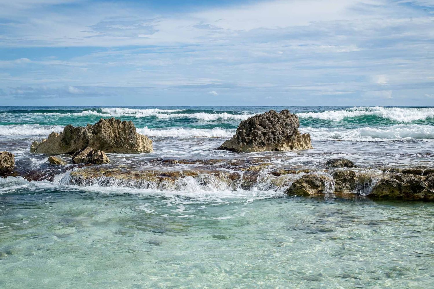 Rocky coastline with turquoise waves crashing against jagged outcrops under a bright blue sky.