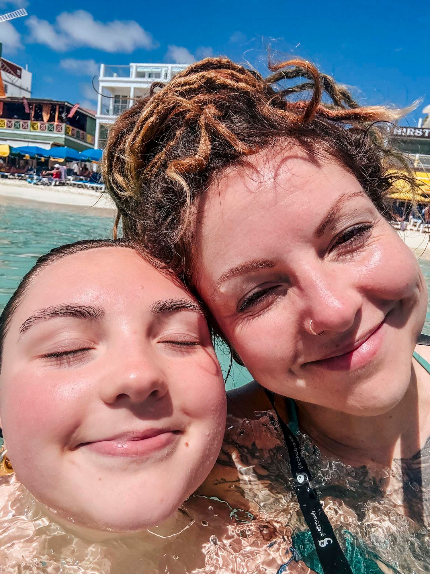Two women smiling in a pool on a sunny day, one with dreadlocks, enjoying a fun outdoor swim.