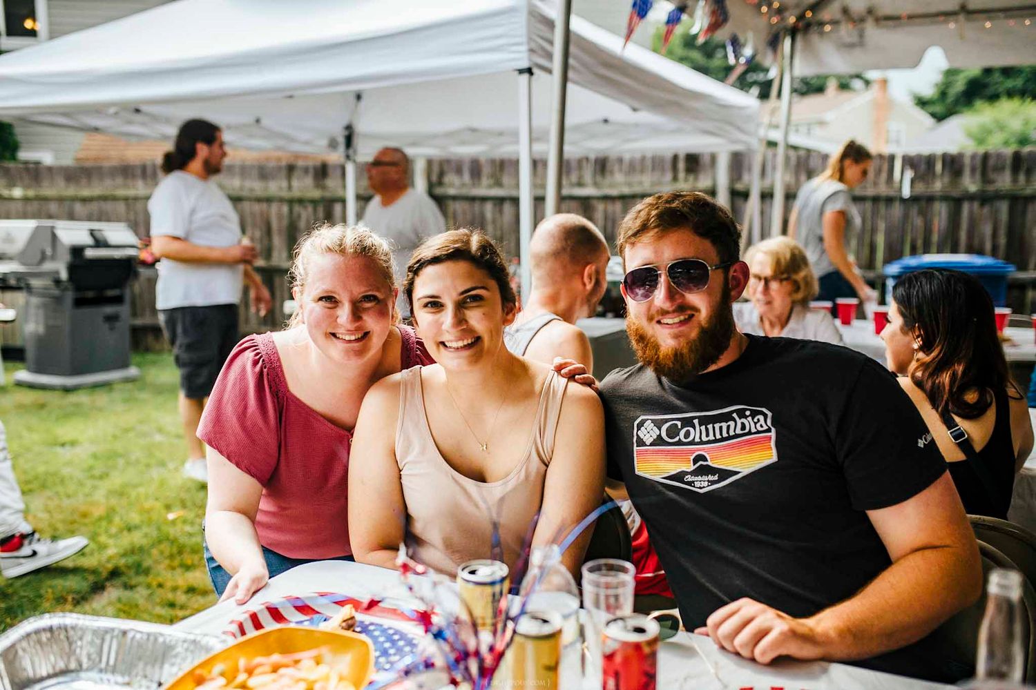 Three friends smiling together at an outdoor backyard barbecue party under a white canopy tent.