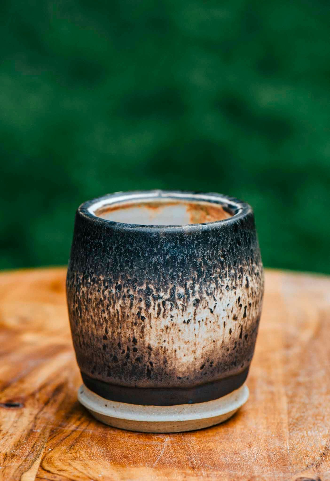 Handcrafted ceramic cup with dark speckled glaze on wooden table, green background.