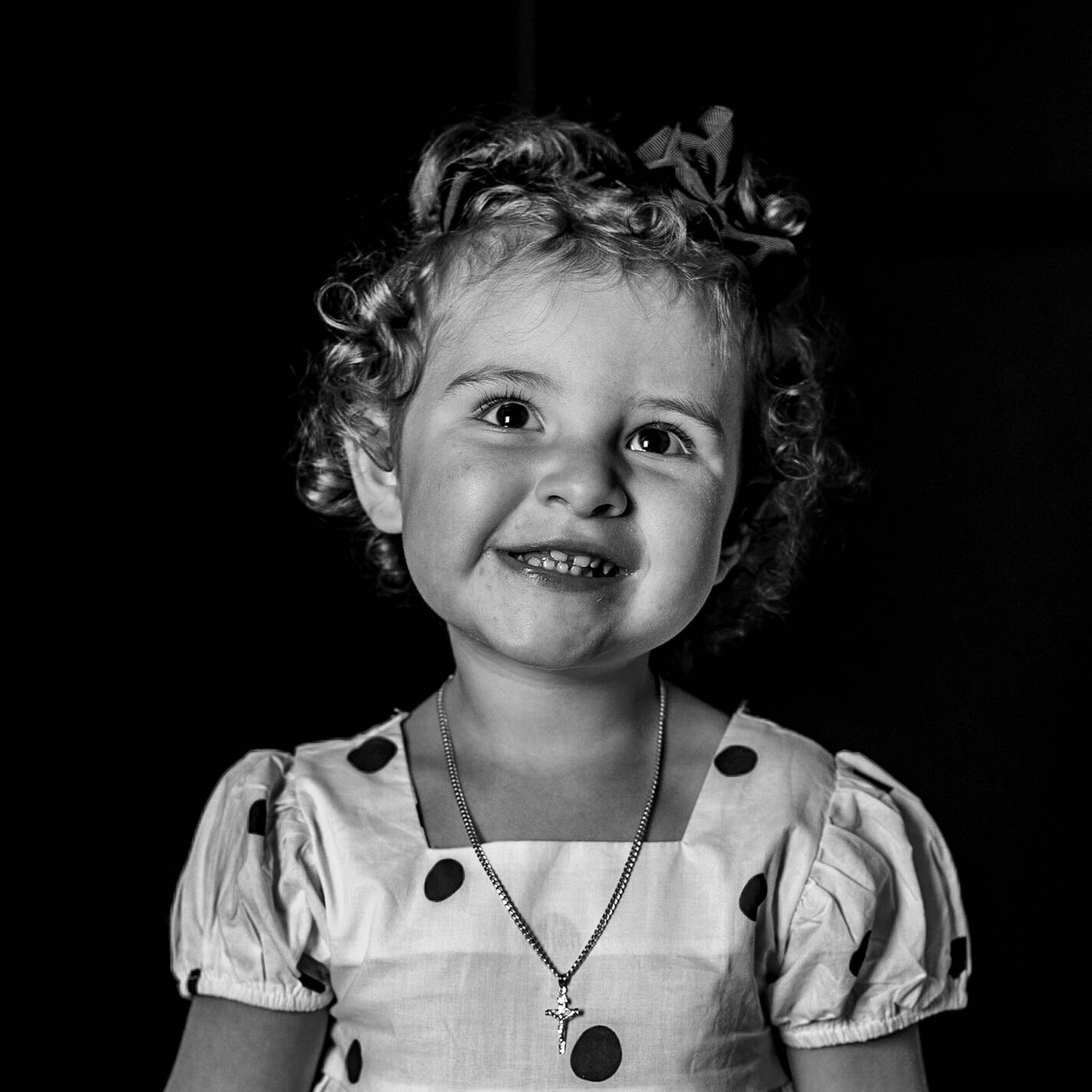 Black and white portrait of a smiling toddler girl with curly hair wearing a polka dot dress and cross necklace.