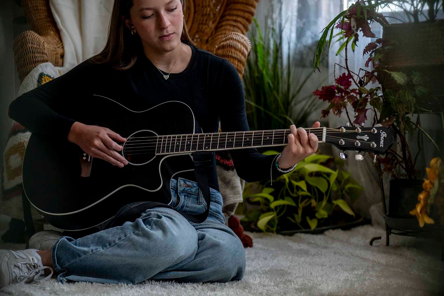 Young woman sitting cross-legged on carpet playing a black acoustic guitar surrounded by indoor plants.