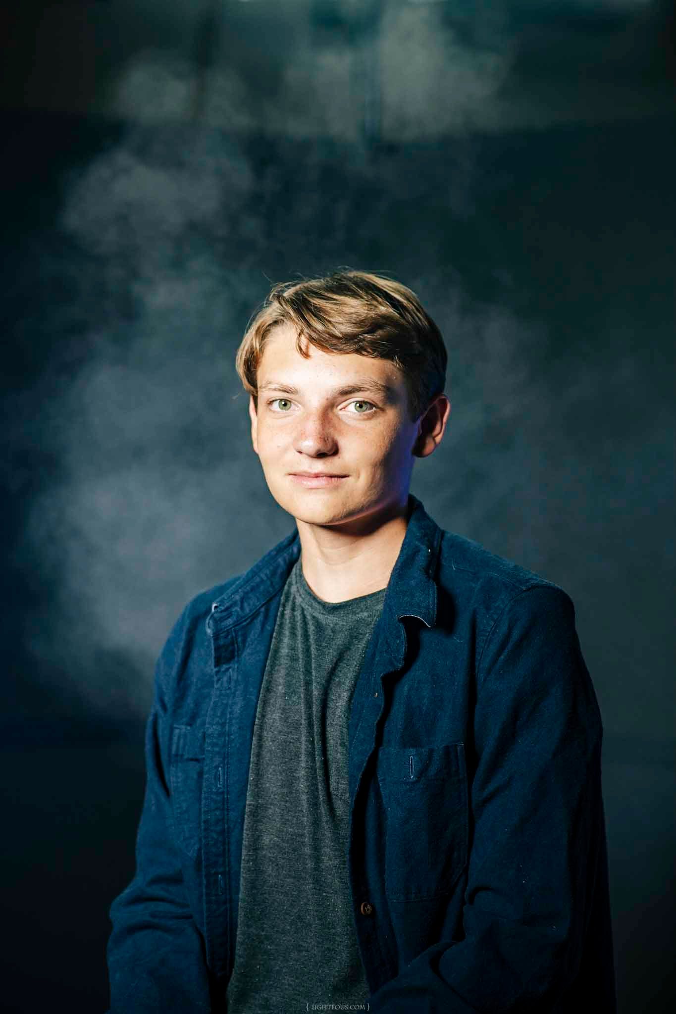 Young man with blonde hair and blue eyes wearing a navy jacket over a gray tee, posing against a dark smoky background.