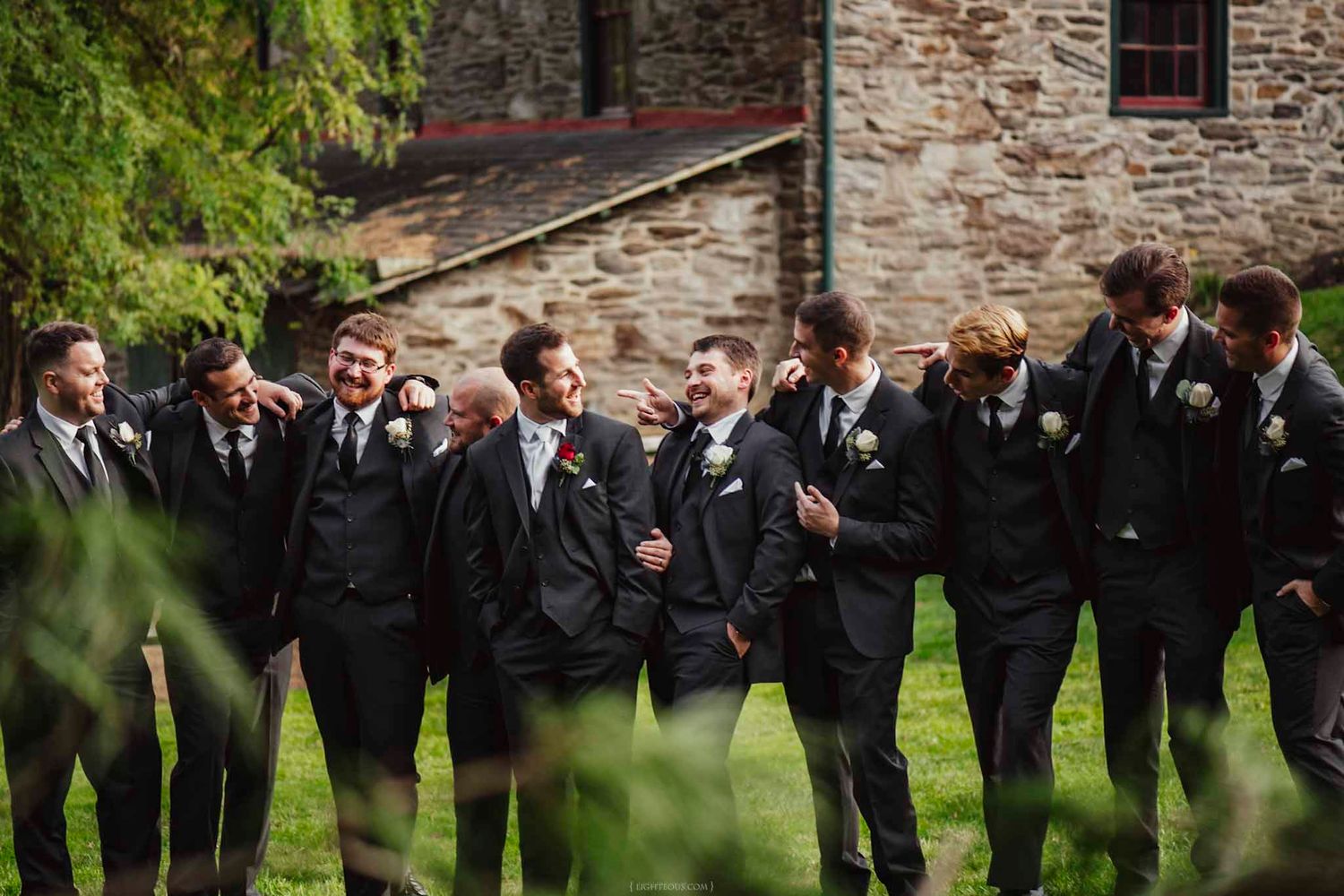 Groomsmen in black suits with boutonnieres laughing together outside a stone building on a wedding day.