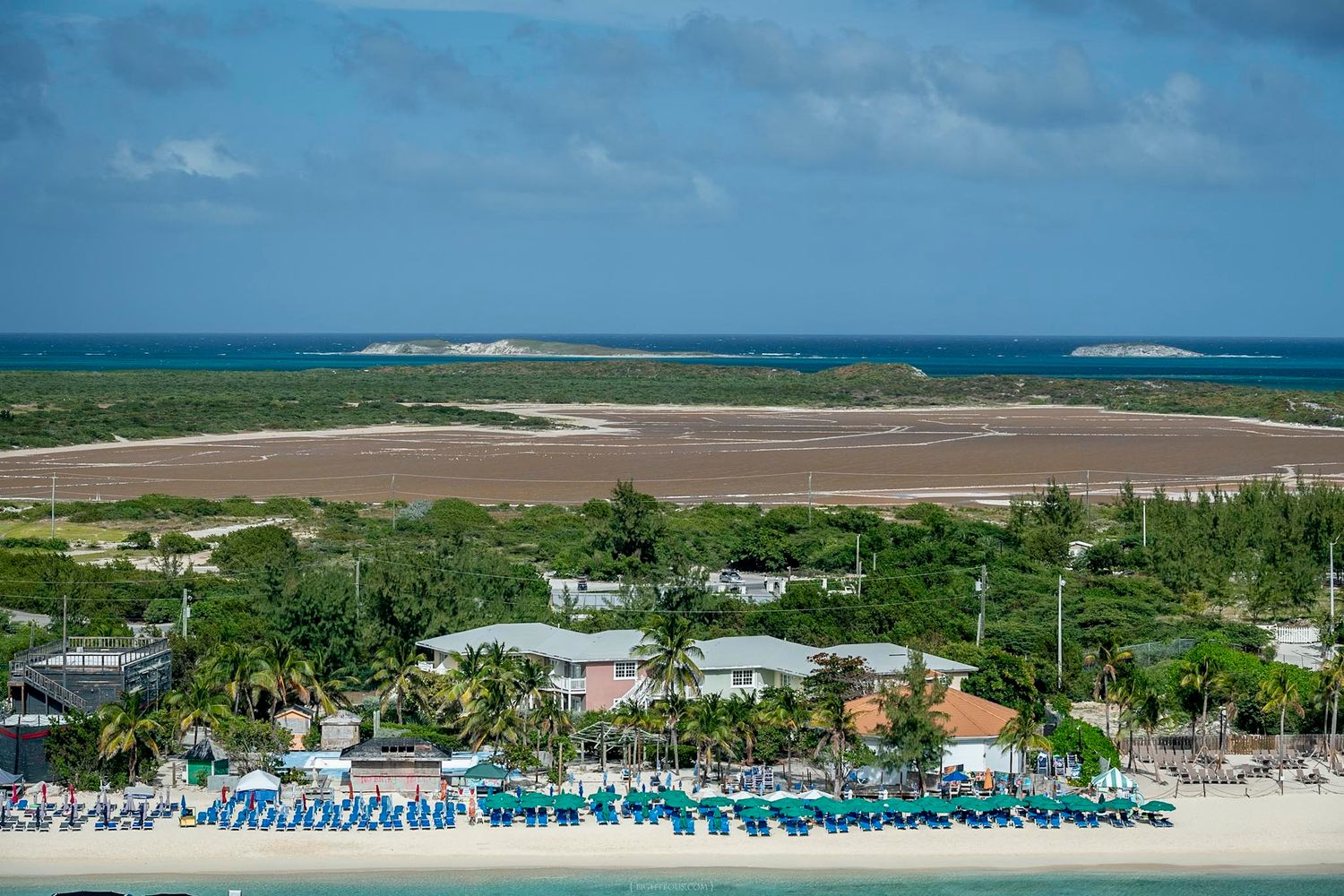 Aerial view of a tropical beach resort with turquoise lounge chairs, lush palms, and a vast salt flat under a partly cloudy sky.