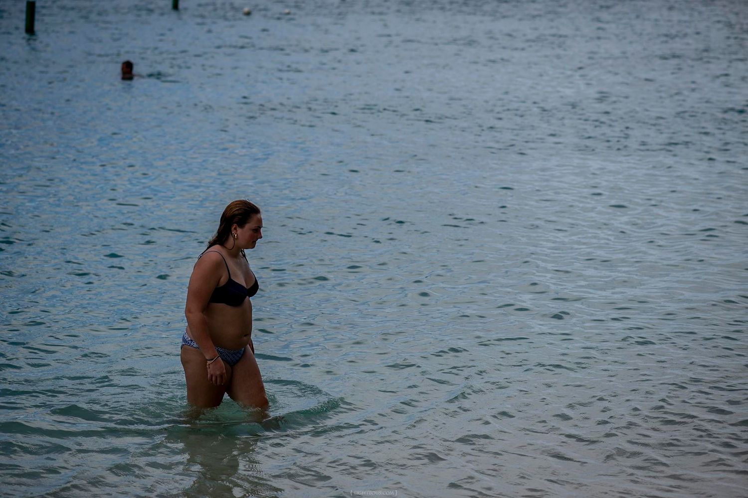 Woman in black bikini wading in calm shallow ocean water near a beach.
