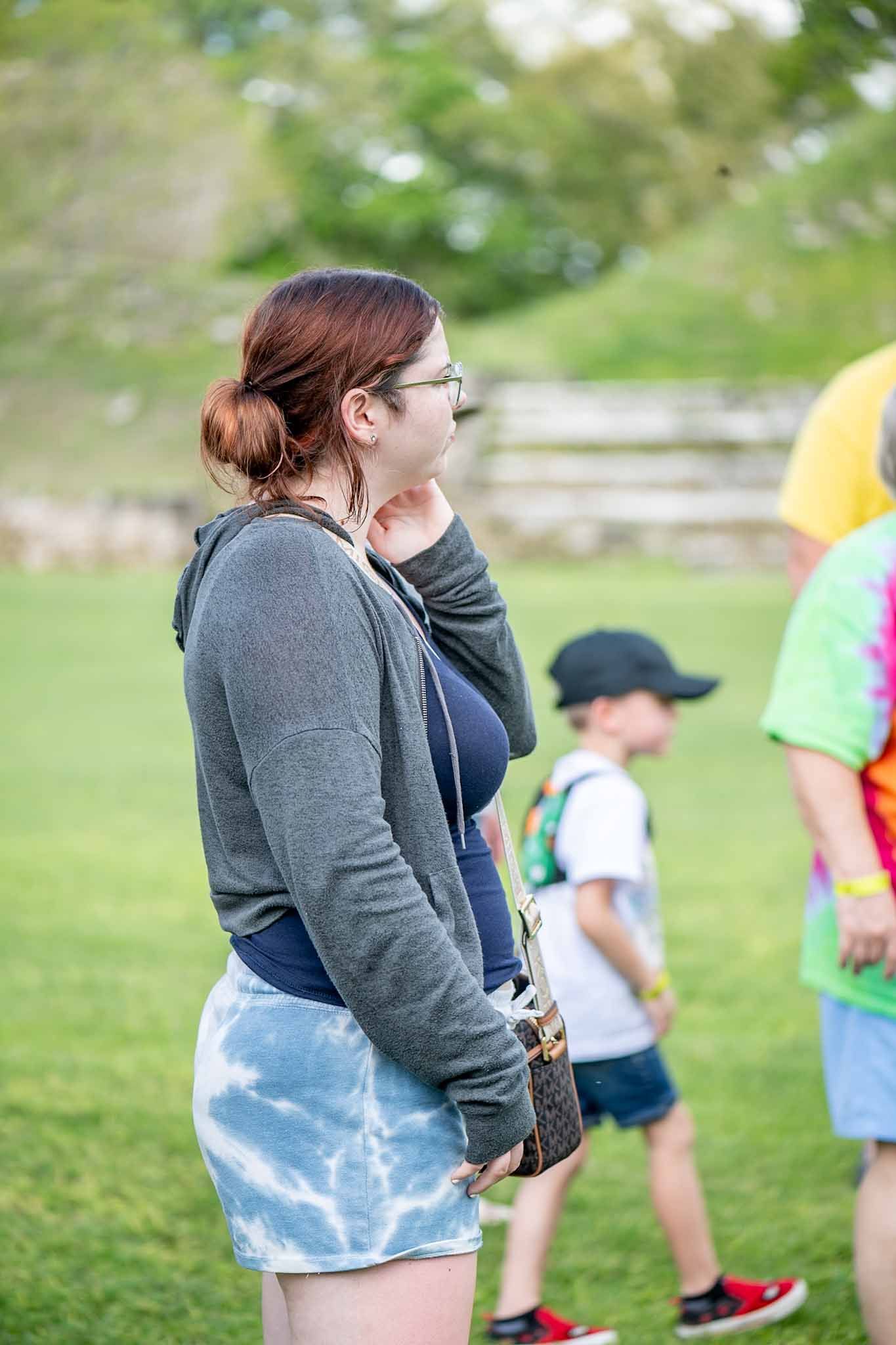 A person wearing a grey sweater and light blue shorts stands outdoors on a grassy field with others in the background.