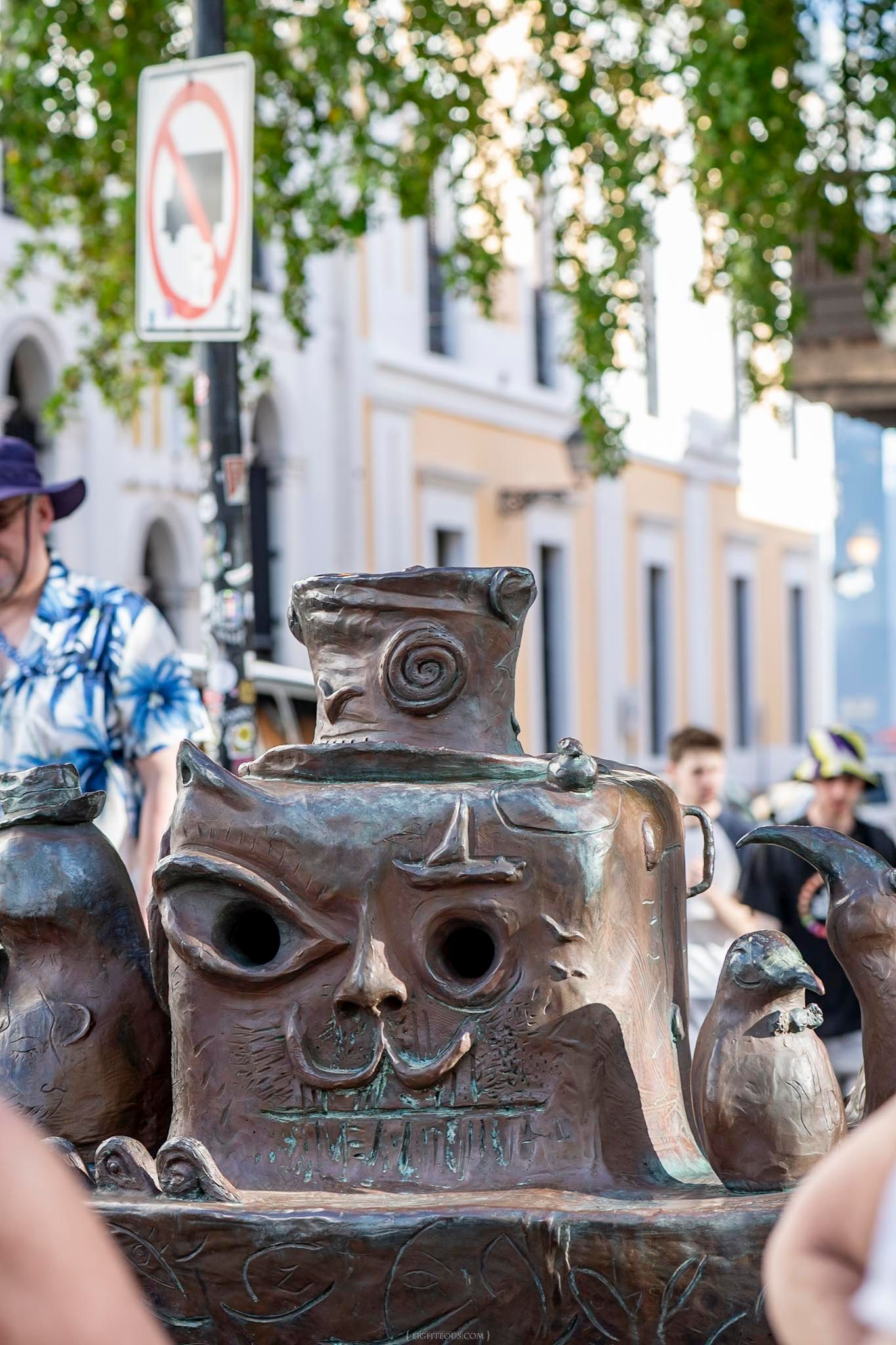 Close-up of a decorative bronze sculpture with a face design displayed outdoors in a busy plaza.