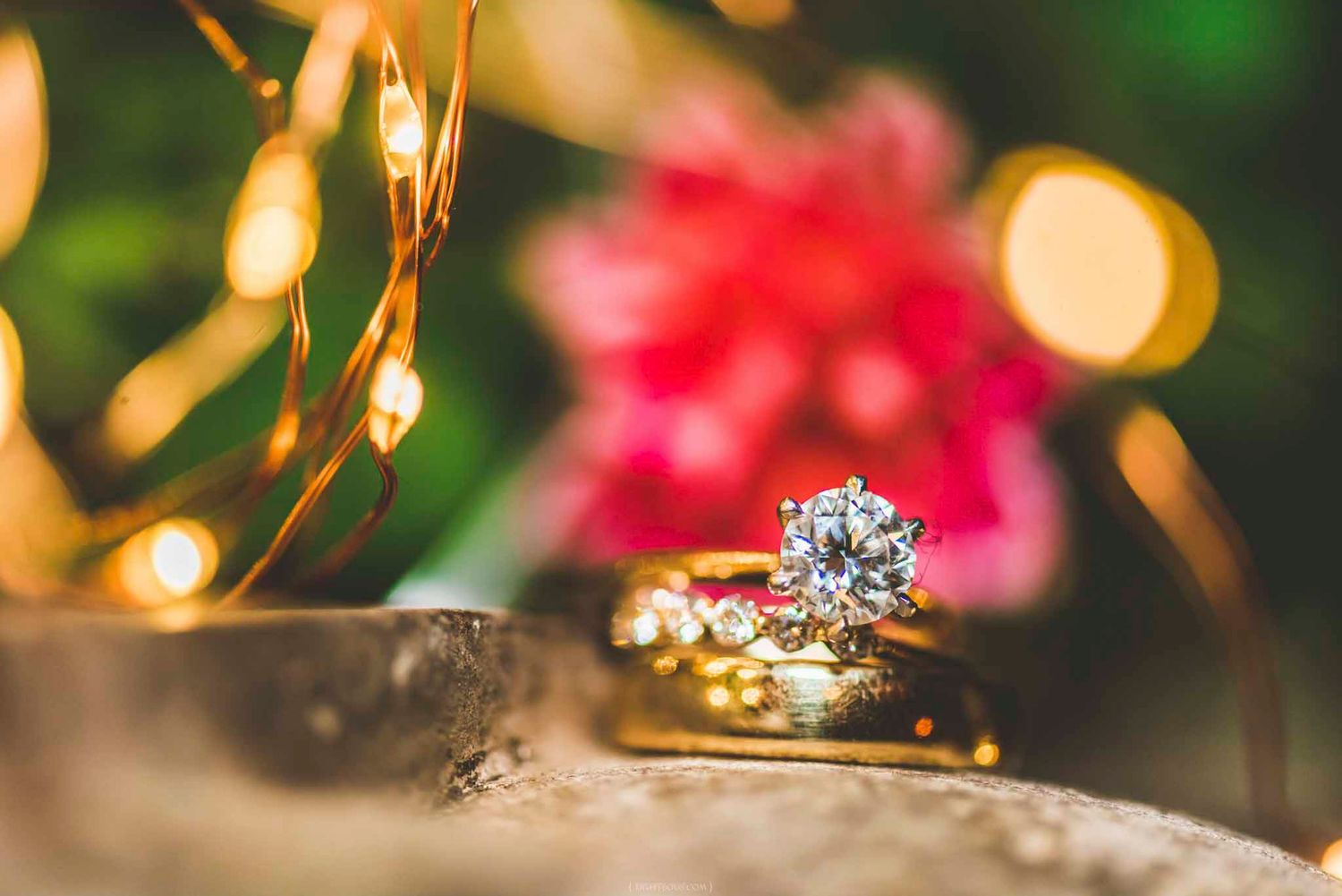 Gold diamond engagement ring and wedding bands stacked on a rustic surface with bokeh floral background.
