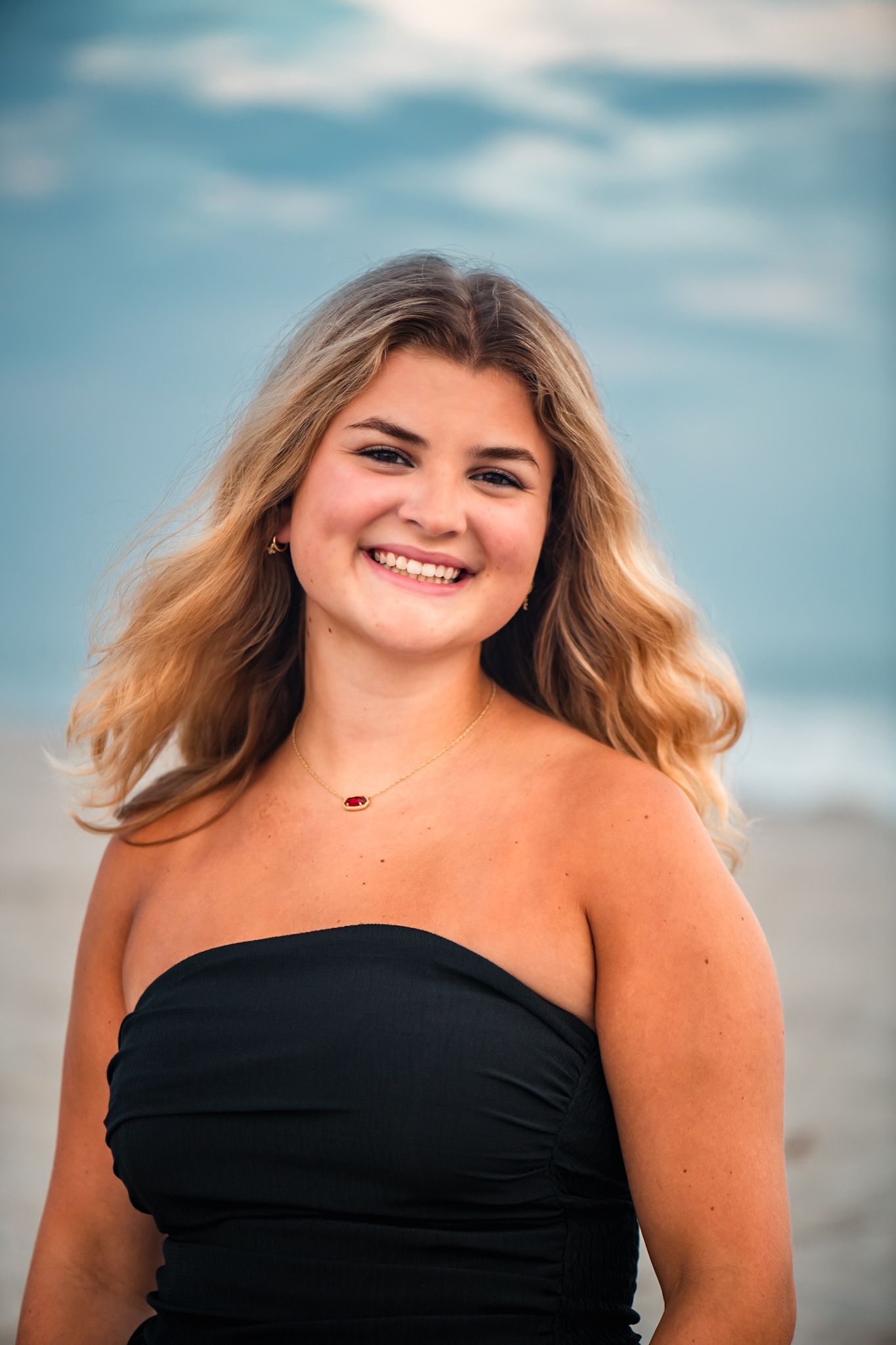Young blonde woman smiling at beach in black strapless top with ocean backdrop.