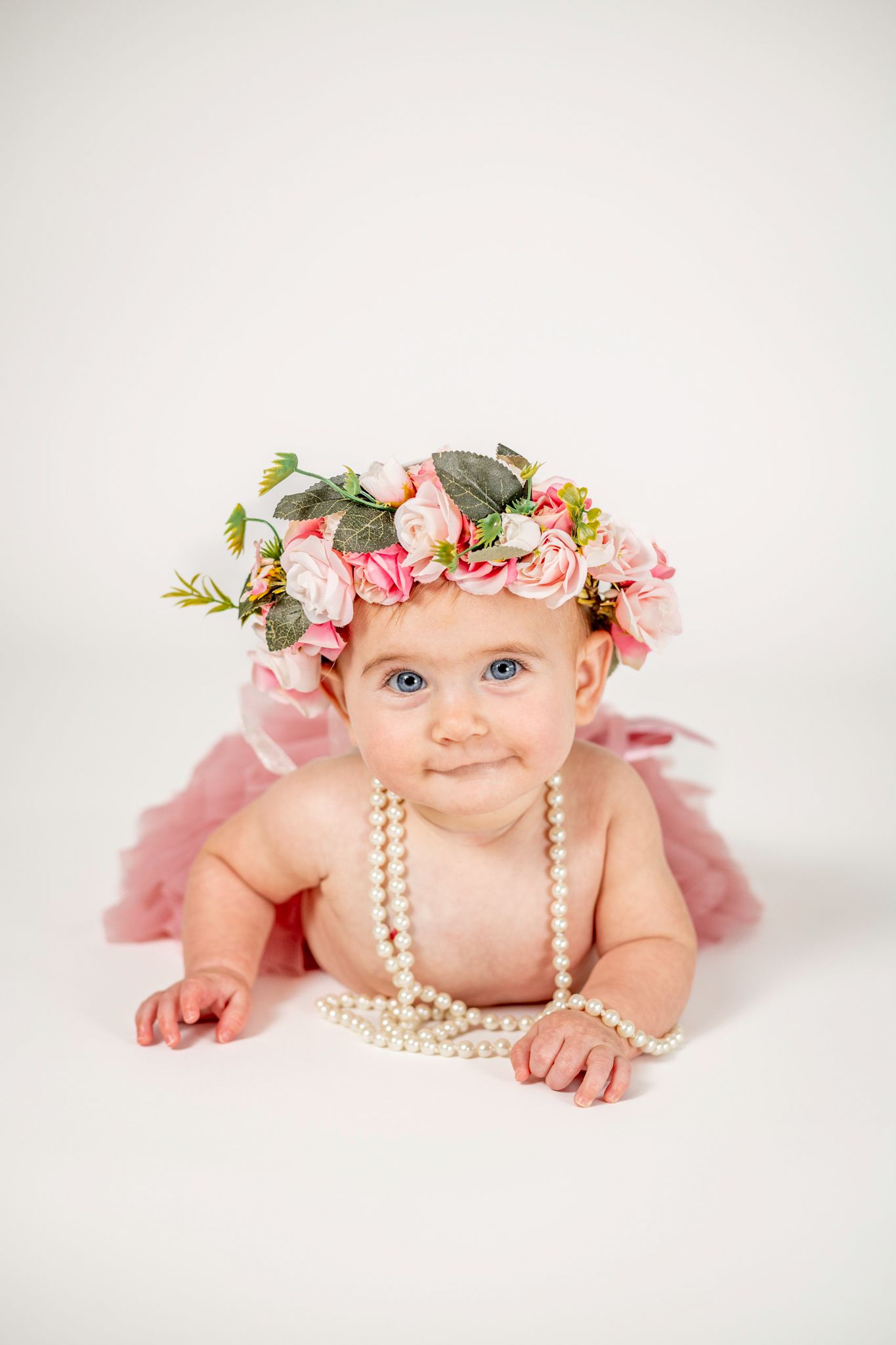Adorable baby girl wearing a pink floral crown and pearl necklace, lying on white background in a pink tutu.