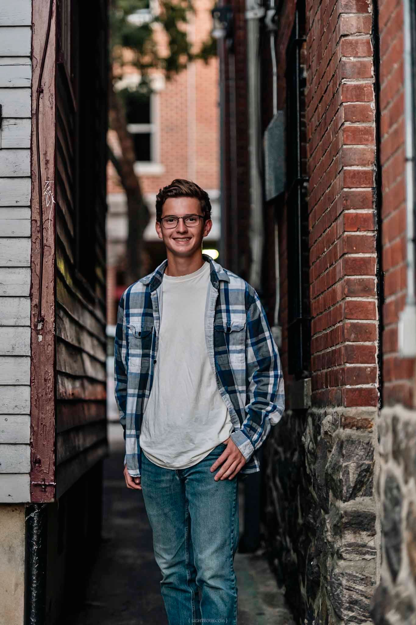 Smiling young man in plaid shirt and jeans standing in urban brick alleyway.