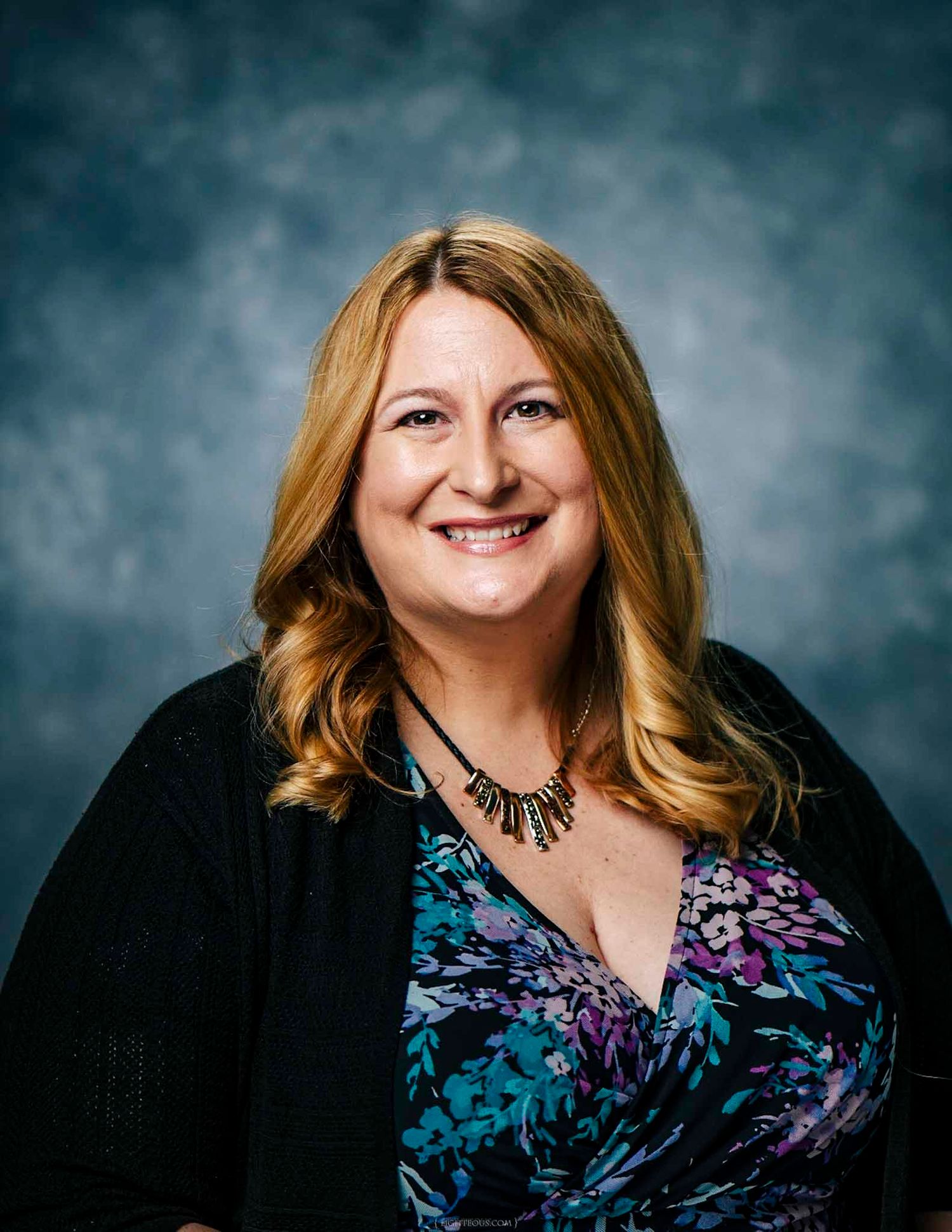 Professional headshot of a smiling woman with blonde hair wearing a floral top and black cardigan against a gray background.