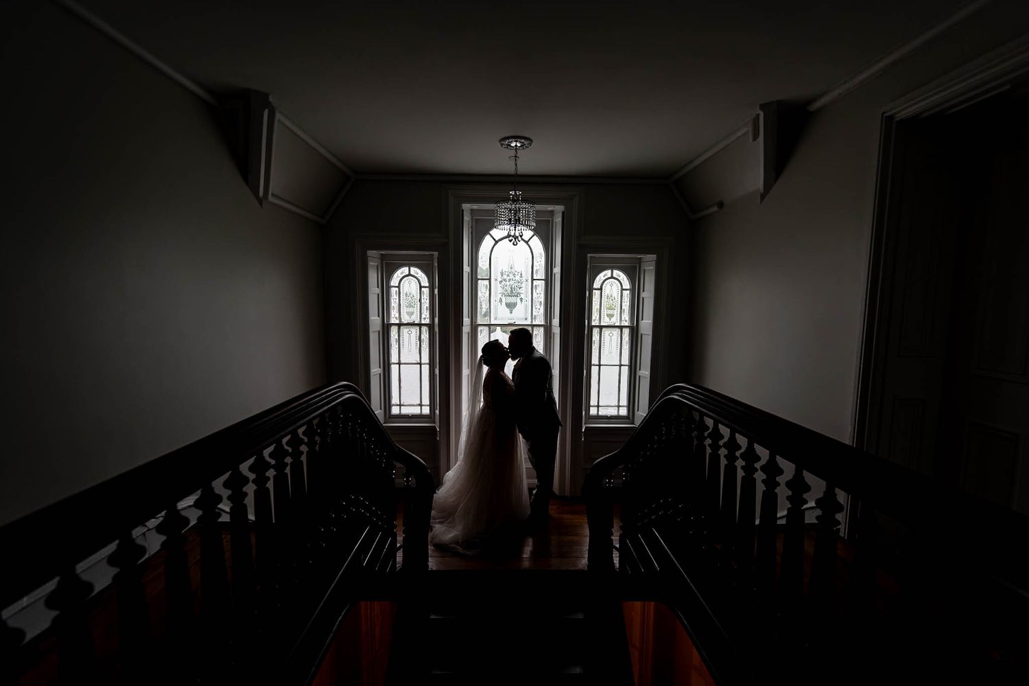 Silhouetted bride and groom share a romantic kiss on a grand staircase with elegant arched windows.