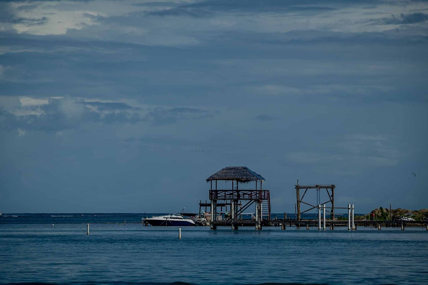 A wooden dock structure with thatched roof stands over calm blue waters against a cloudy sky in the Caribbean.