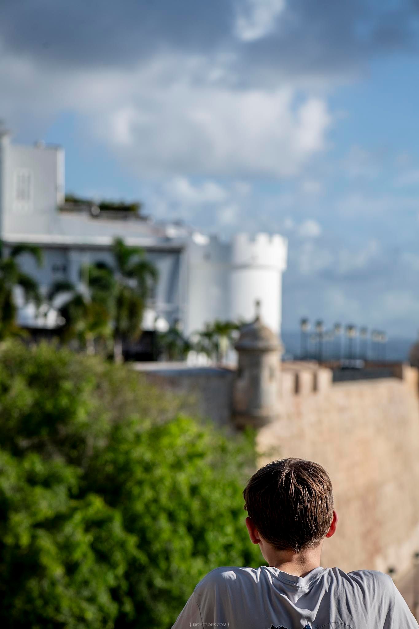 Young man viewed from behind gazing at historic stone fortress walls and white buildings under a partly cloudy sky.