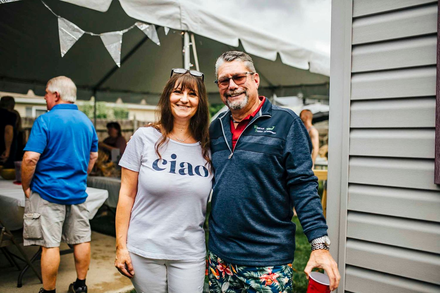 A smiling couple poses together outdoors at a casual event, woman wearing a Ciao shirt, man in navy jacket with colorful shorts.