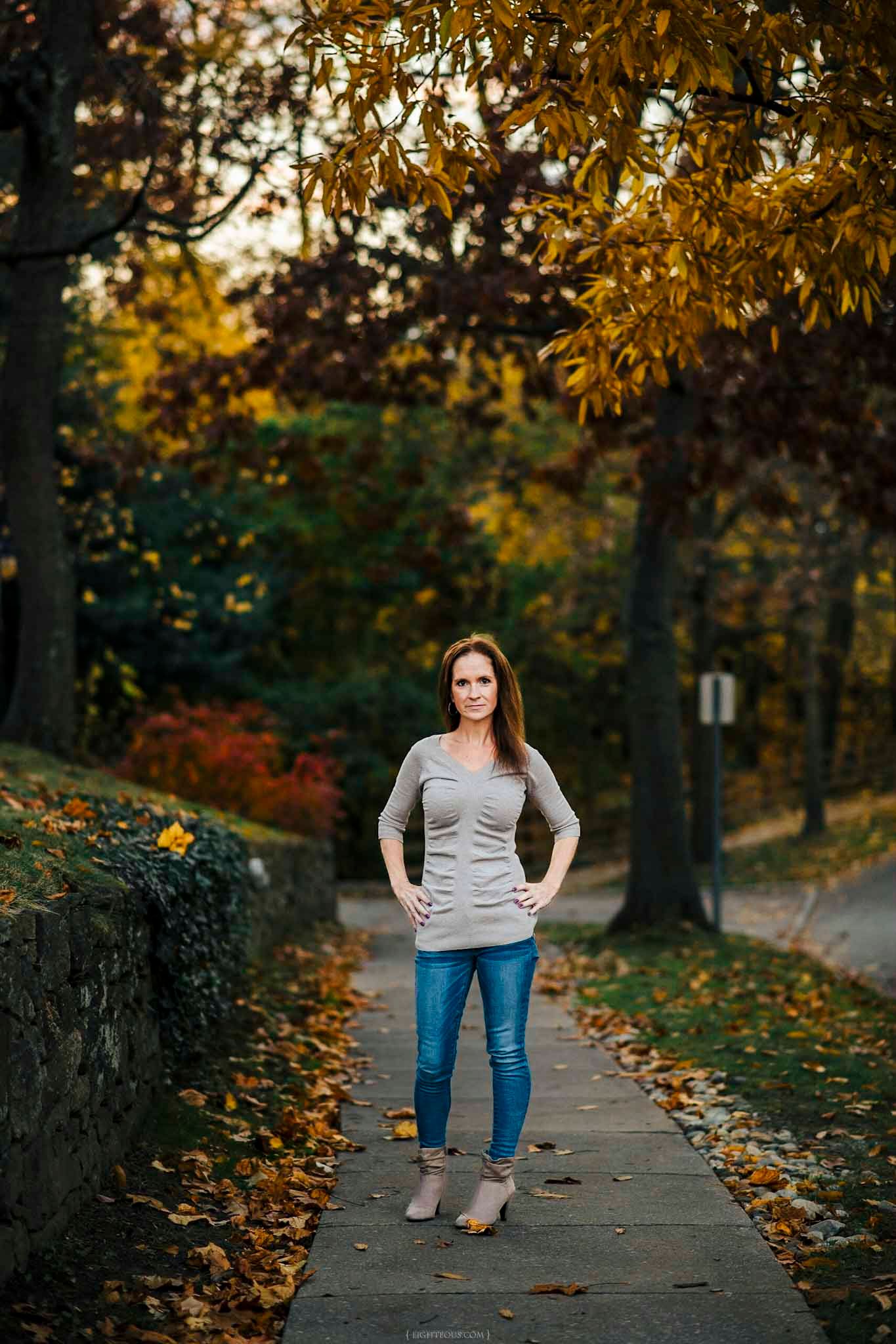 Woman in gray top and jeans stands on a tree-lined path surrounded by colorful autumn foliage.