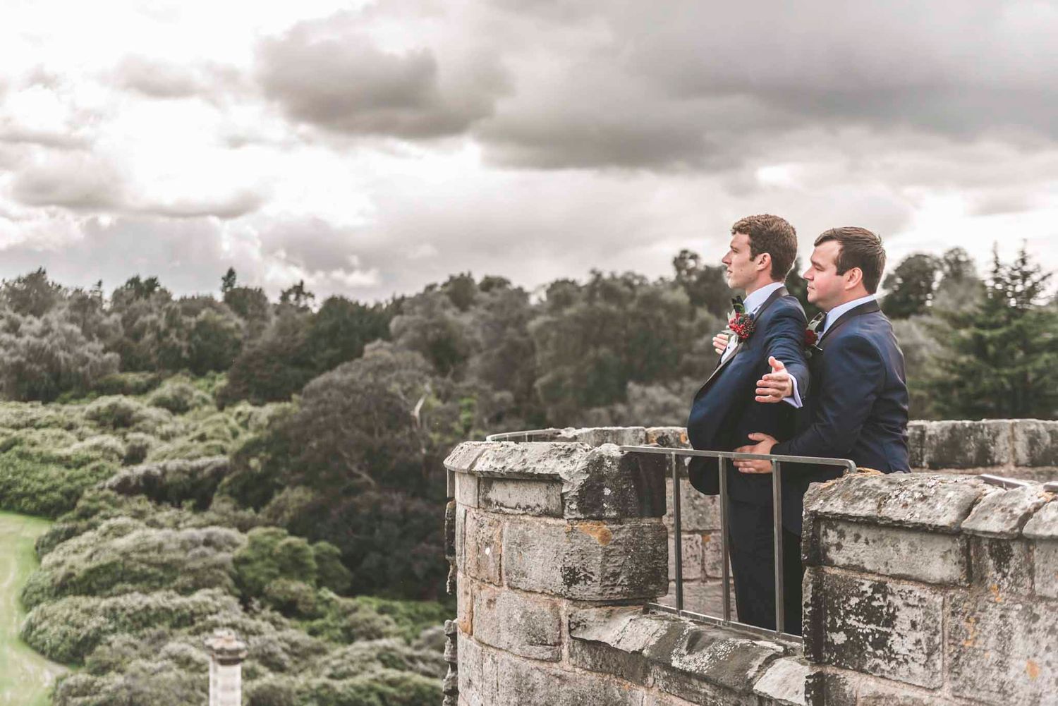 Two grooms in navy suits stand on a castle balcony overlooking lush green gardens under dramatic cloudy skies.
