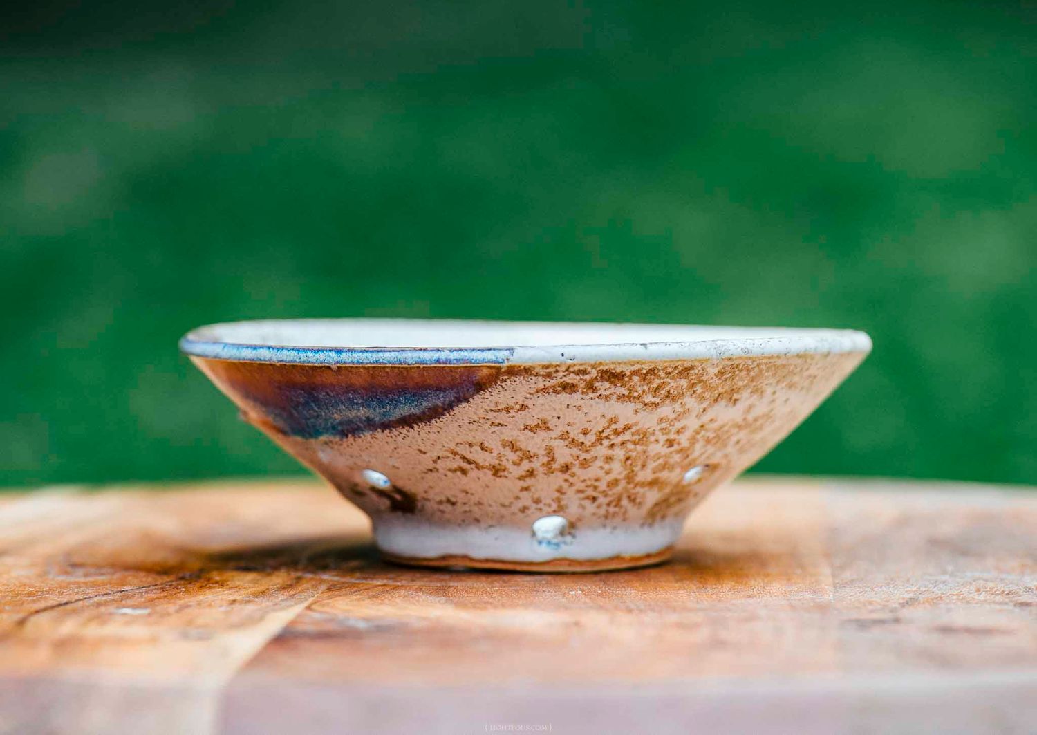 Handcrafted ceramic bowl with brown and cream glaze resting on a wooden surface against a green background.