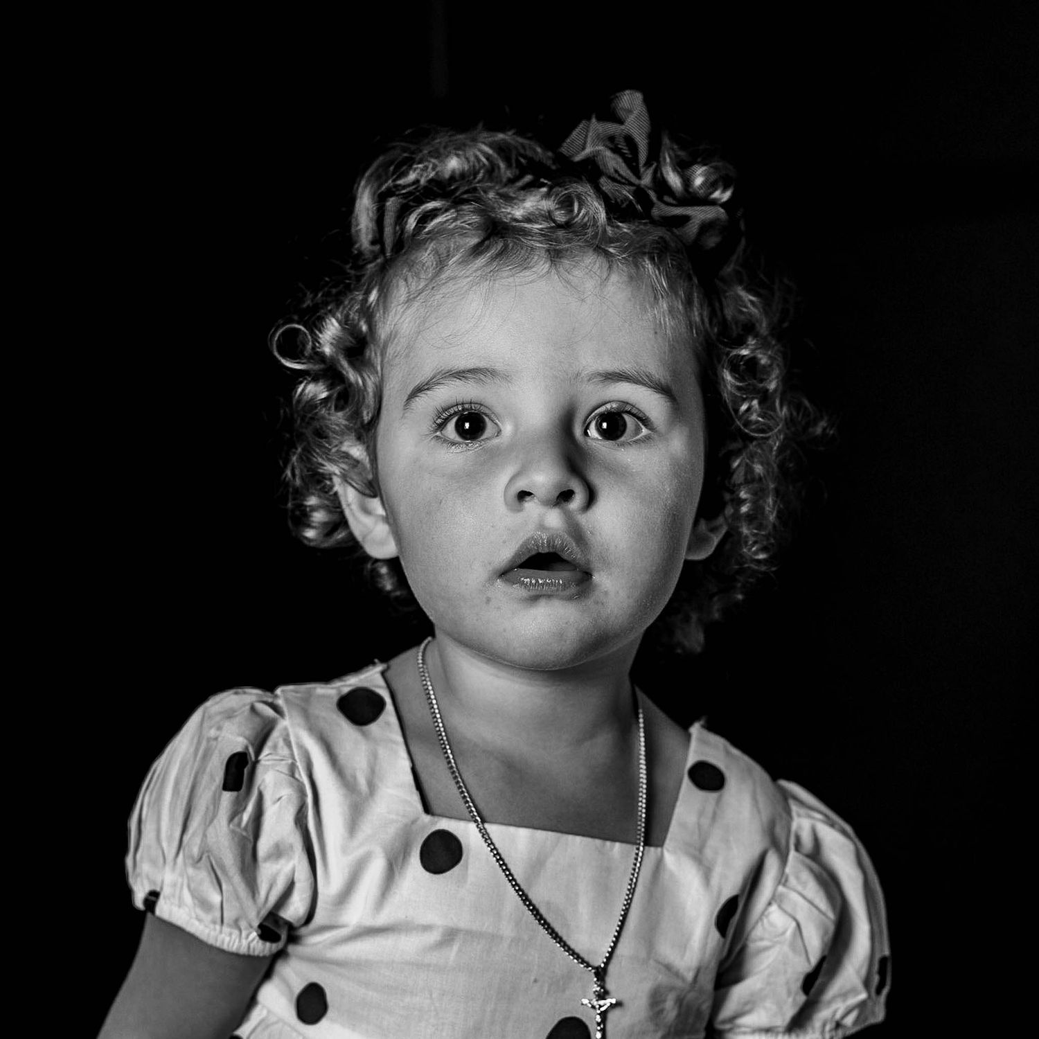 Black and white portrait of a curly-haired toddler girl in a polka dot dress with a necklace, looking surprised.