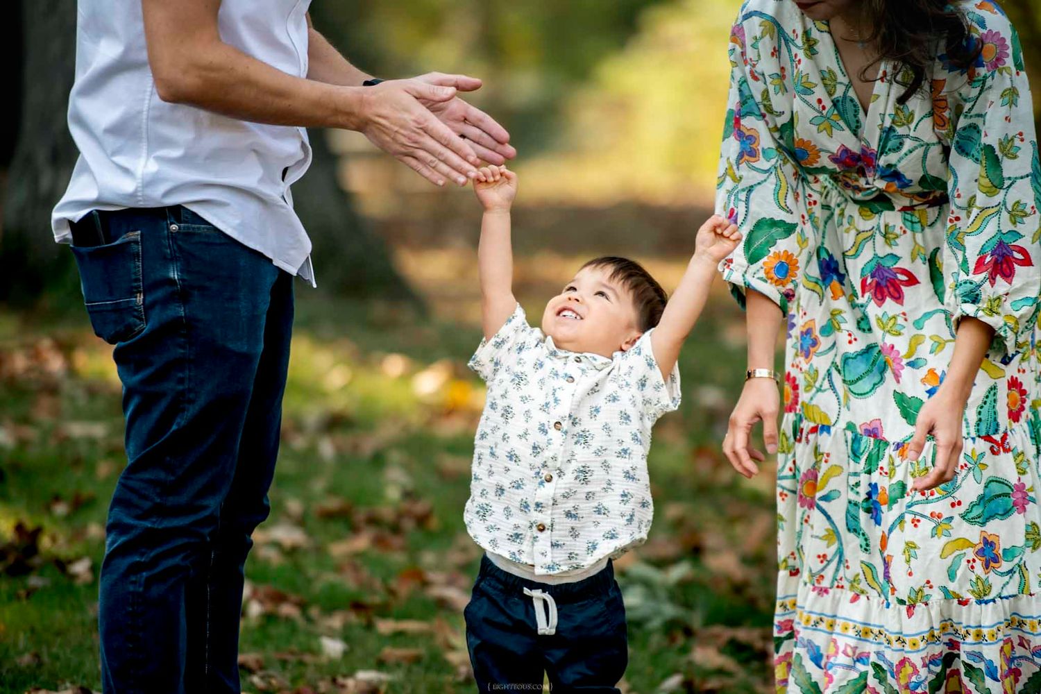 A joyful toddler laughs while holding hands with parents outdoors in a fall park setting.