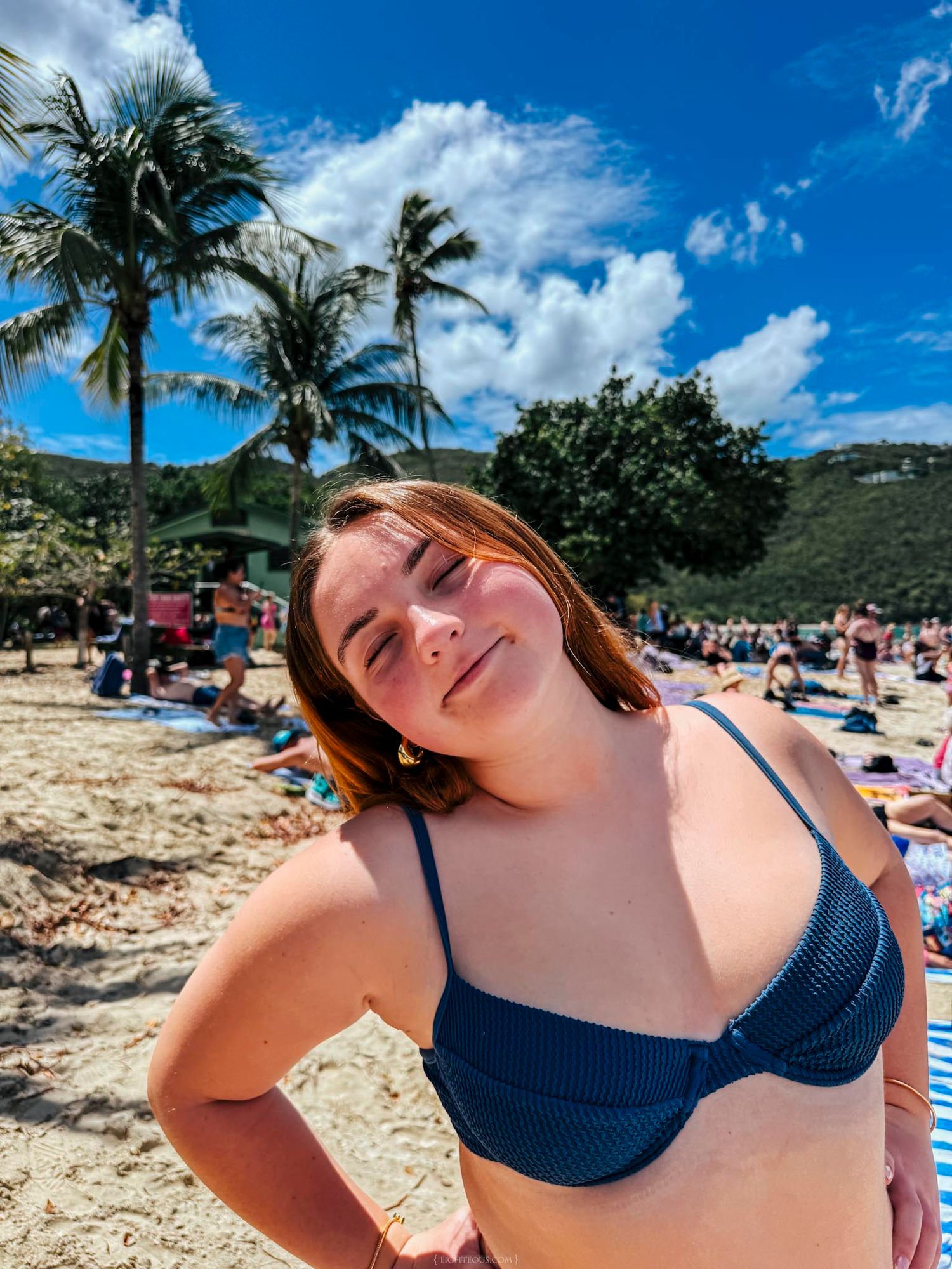 Young woman in teal bikini smiling on sunny tropical beach with palm trees and blue sky.
