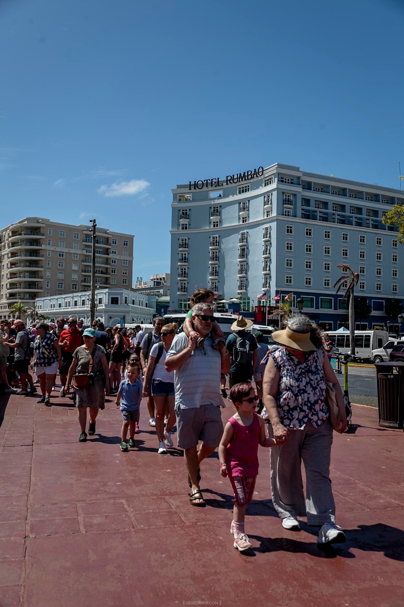 Crowds of tourists walk along a waterfront promenade with Hotel Rumbo visible in the background under a clear blue sky.