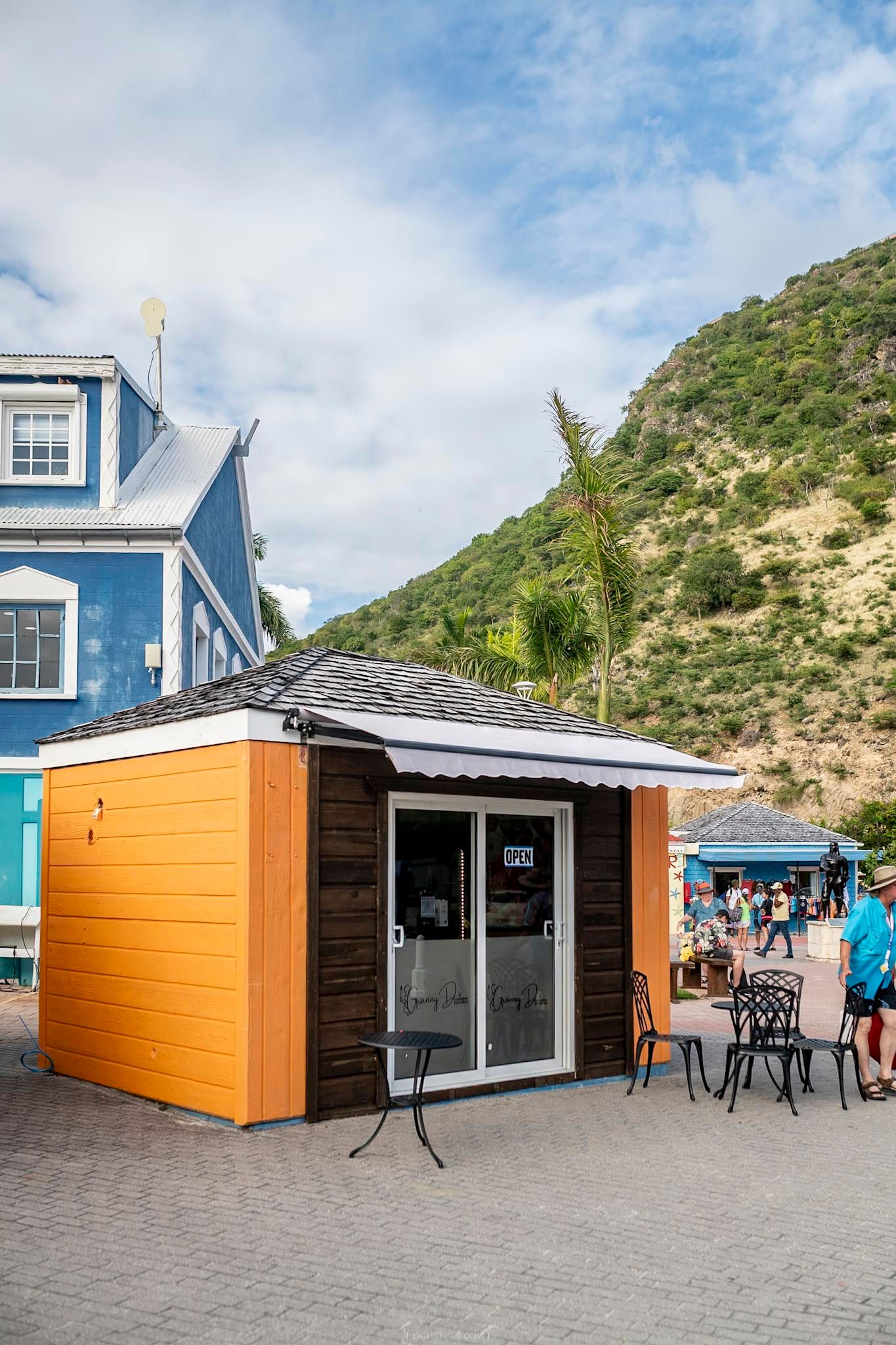 Small orange and brown beach kiosk with open sign near a blue building and green hillside backdrop.