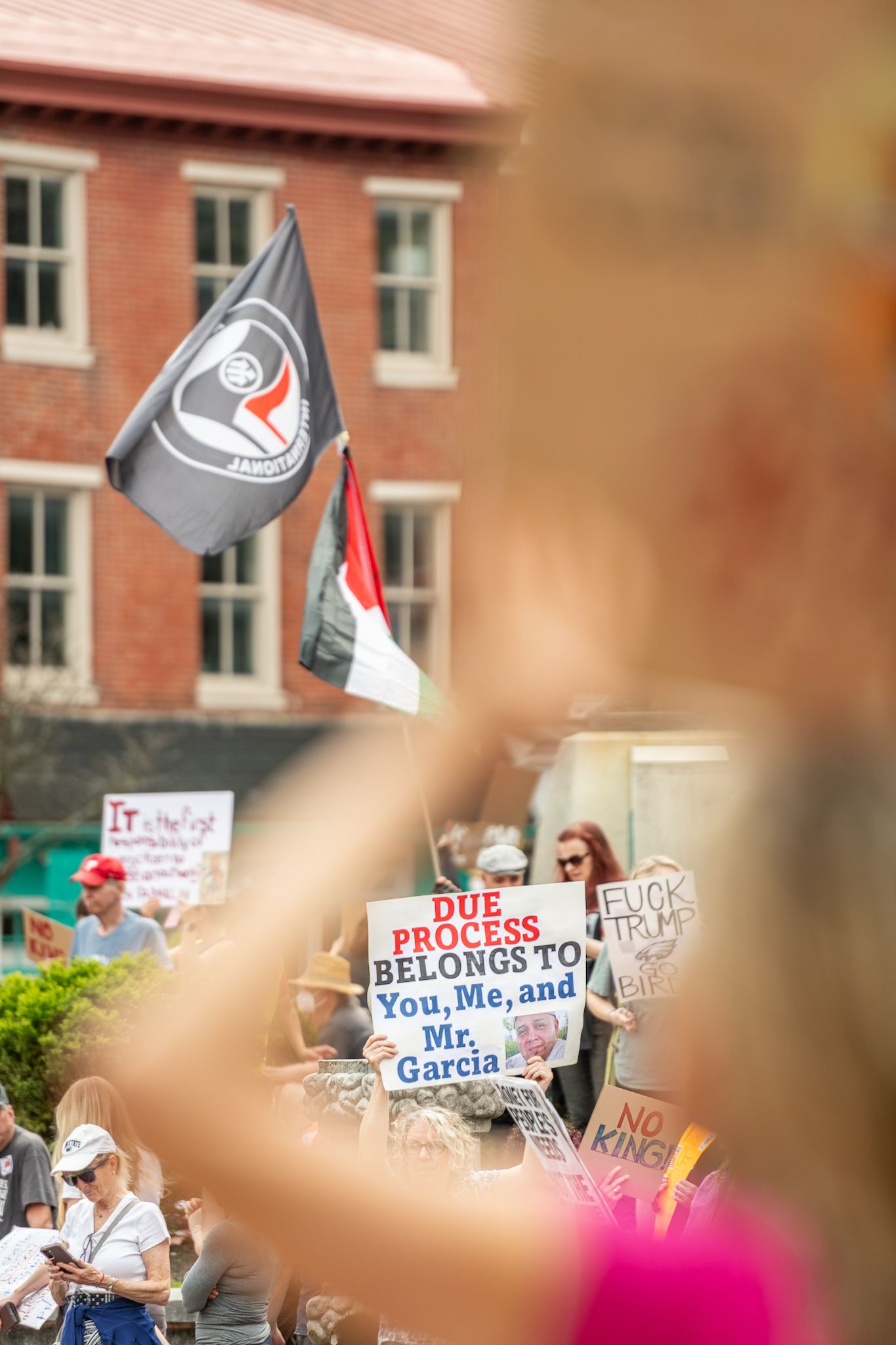 Political protest with various signs and flags visible against brick building backdrop.