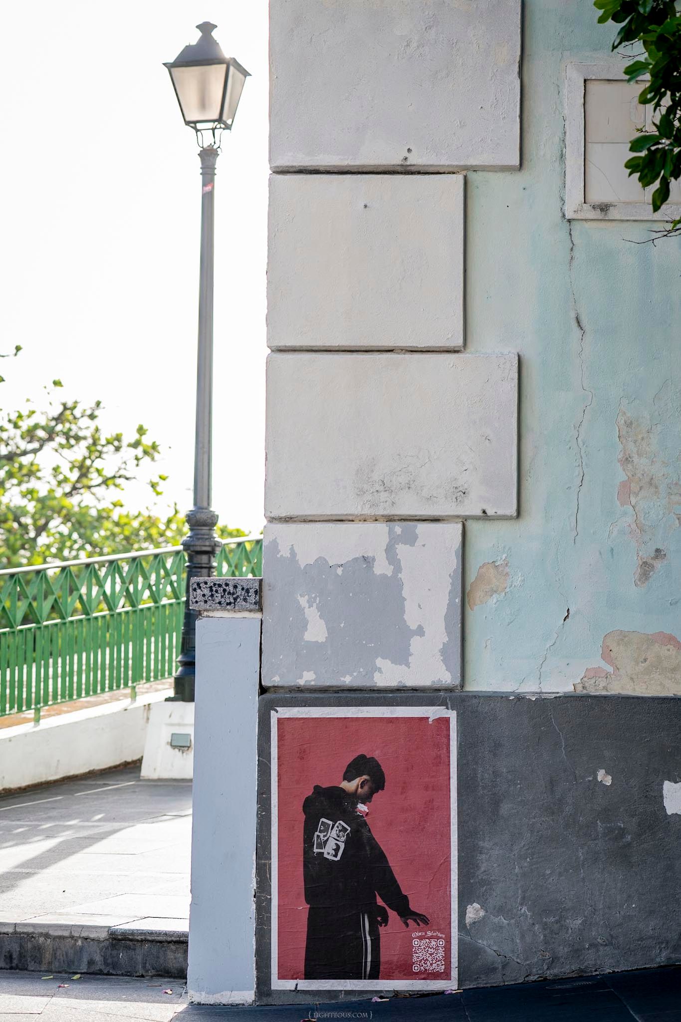 Street art poster of a figure in black on a weathered building corner with a vintage lamp post in an urban setting.