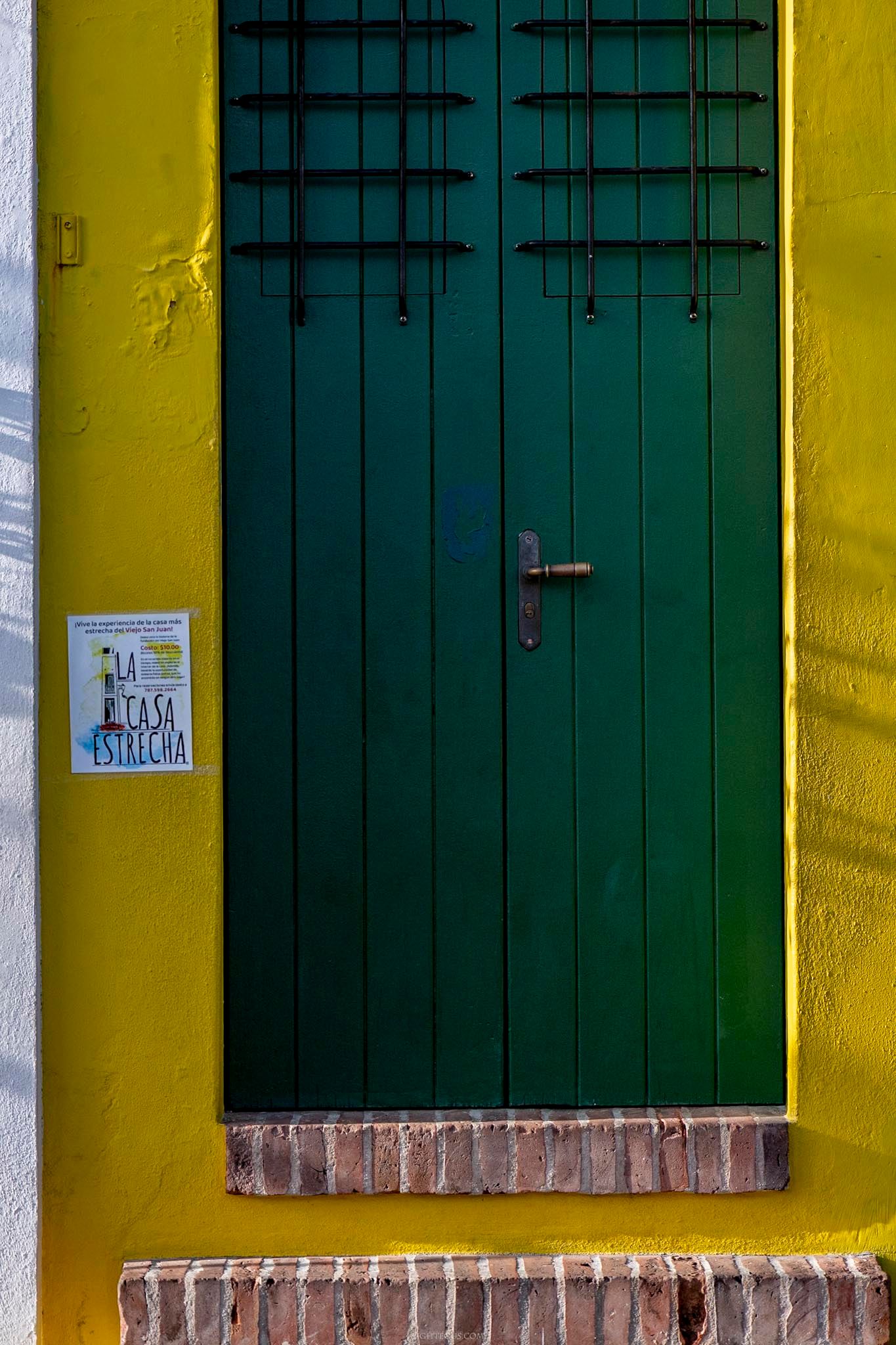 Green wooden door with iron bars set in a bright yellow wall with brick threshold in Cartagena, Colombia.