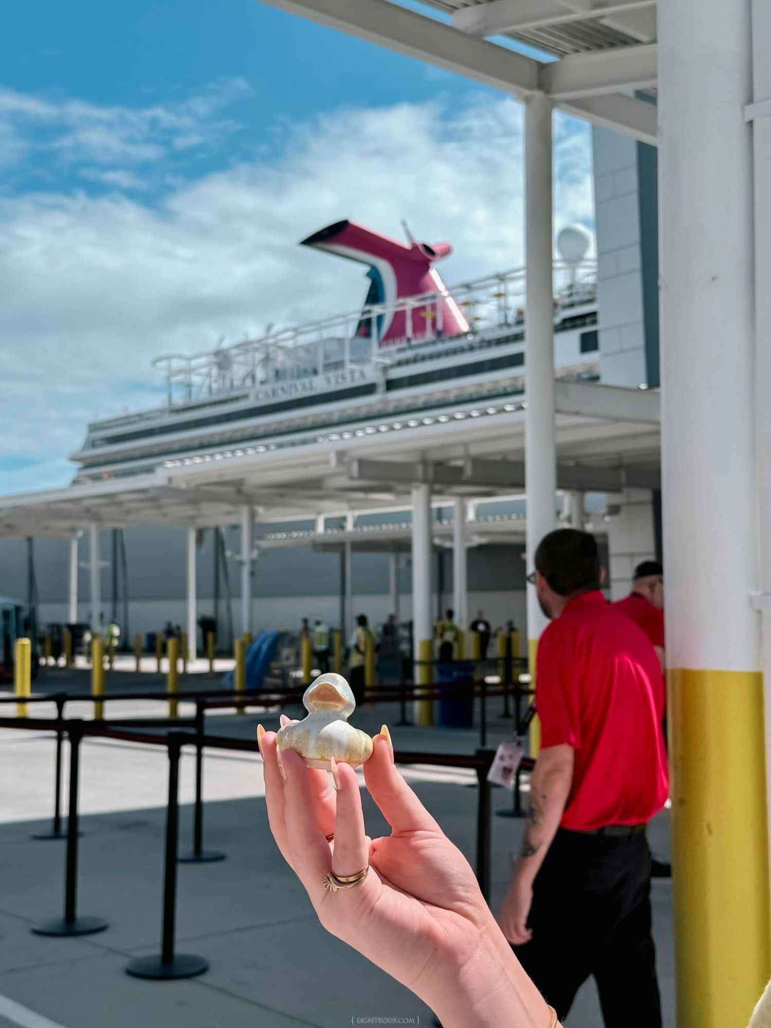 Hand holding a small figurine at a Carnival cruise ship terminal with the iconic red funnel visible in the background.