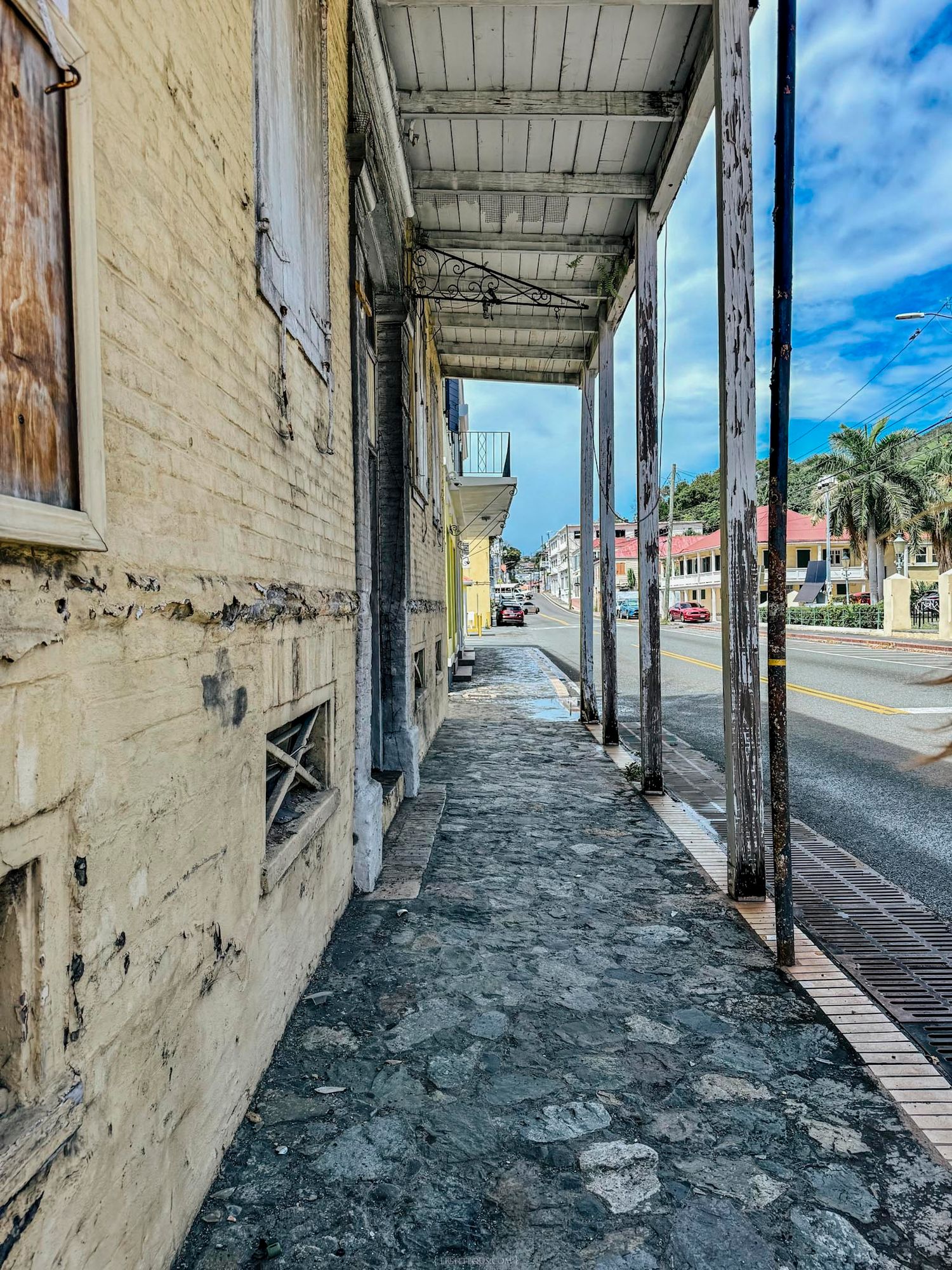 Historic cobblestone sidewalk beneath a weathered colonial arcade in a Caribbean town with palm trees and blue sky.