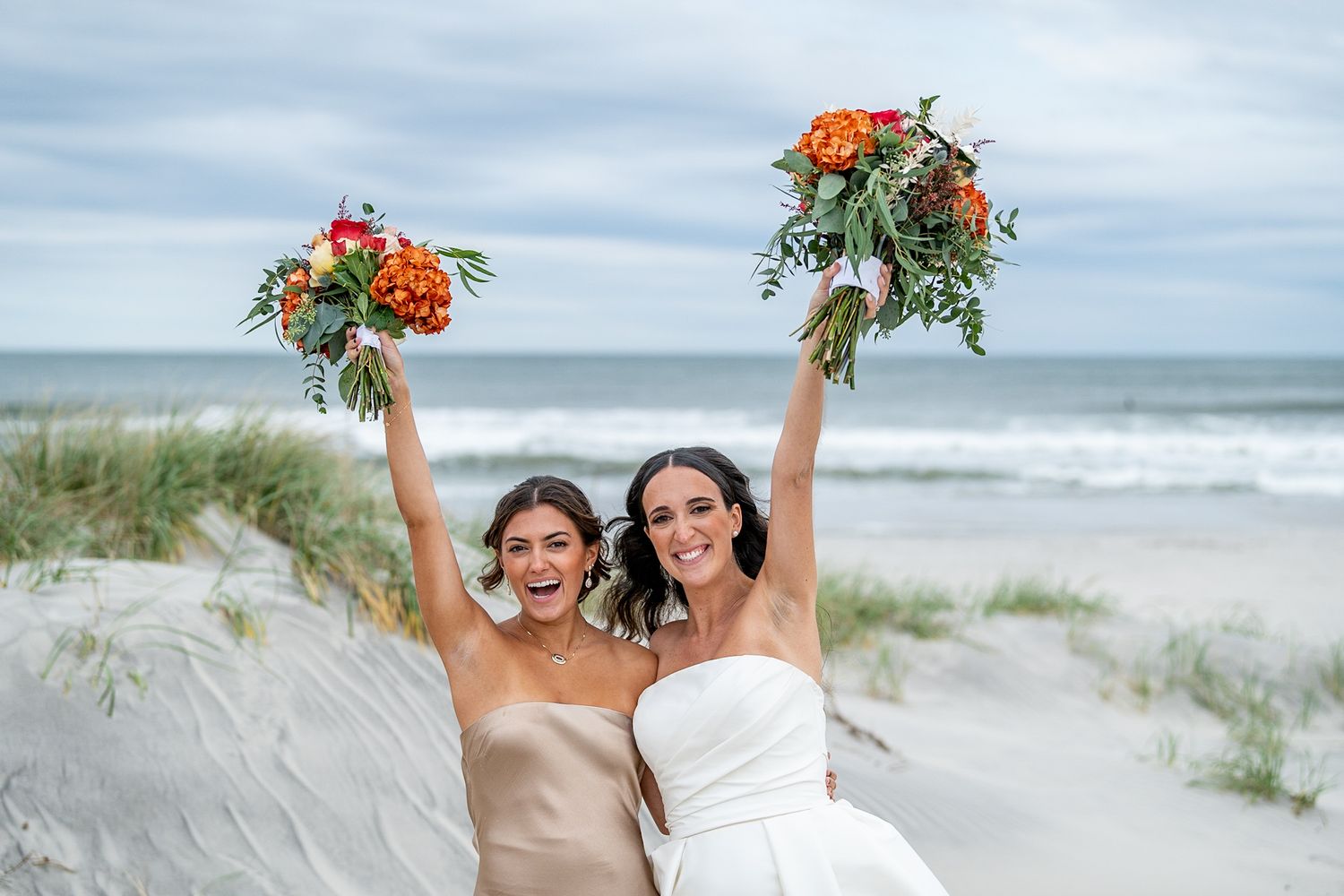 Two brides celebrate on a beach, raising colorful bouquets, one in white gown, one in champagne dress.