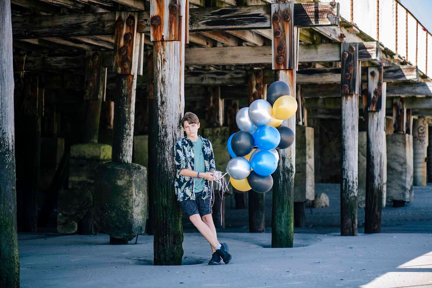 Person holding colorful balloons under a pier with wooden pillars on a sunny day.