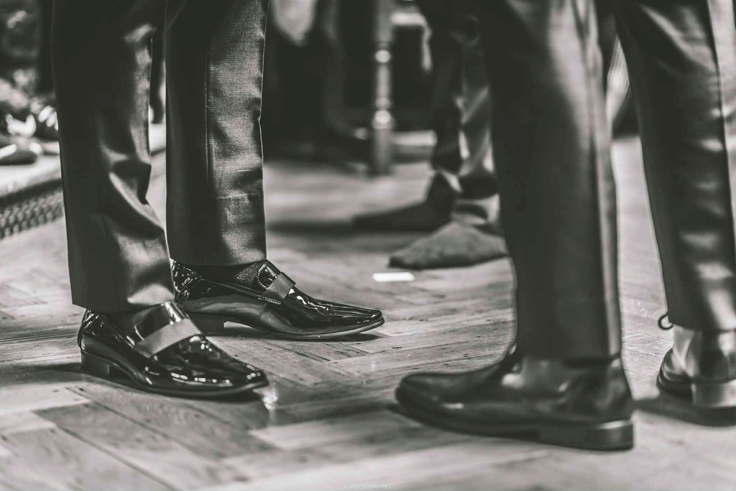 Black and white photo of men in formal dress shoes and suit trousers standing on a wooden floor.
