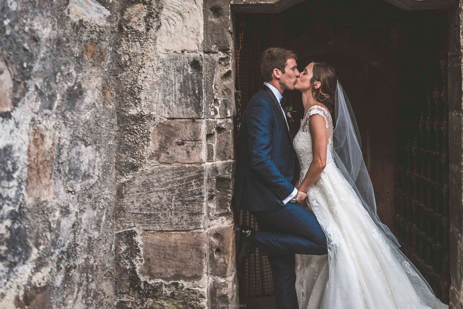 A bride and groom share a romantic kiss in a rustic stone doorway, the bride wearing a white gown with veil.