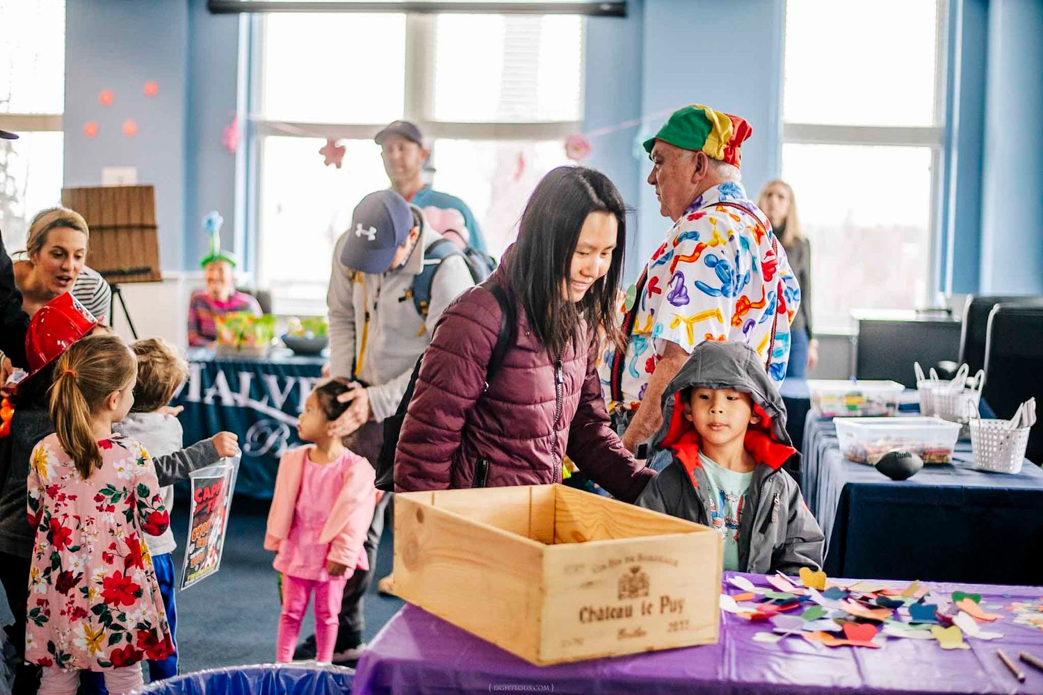 Families enjoy a colorful craft activity booth at an indoor children's event with a clown entertainer nearby.