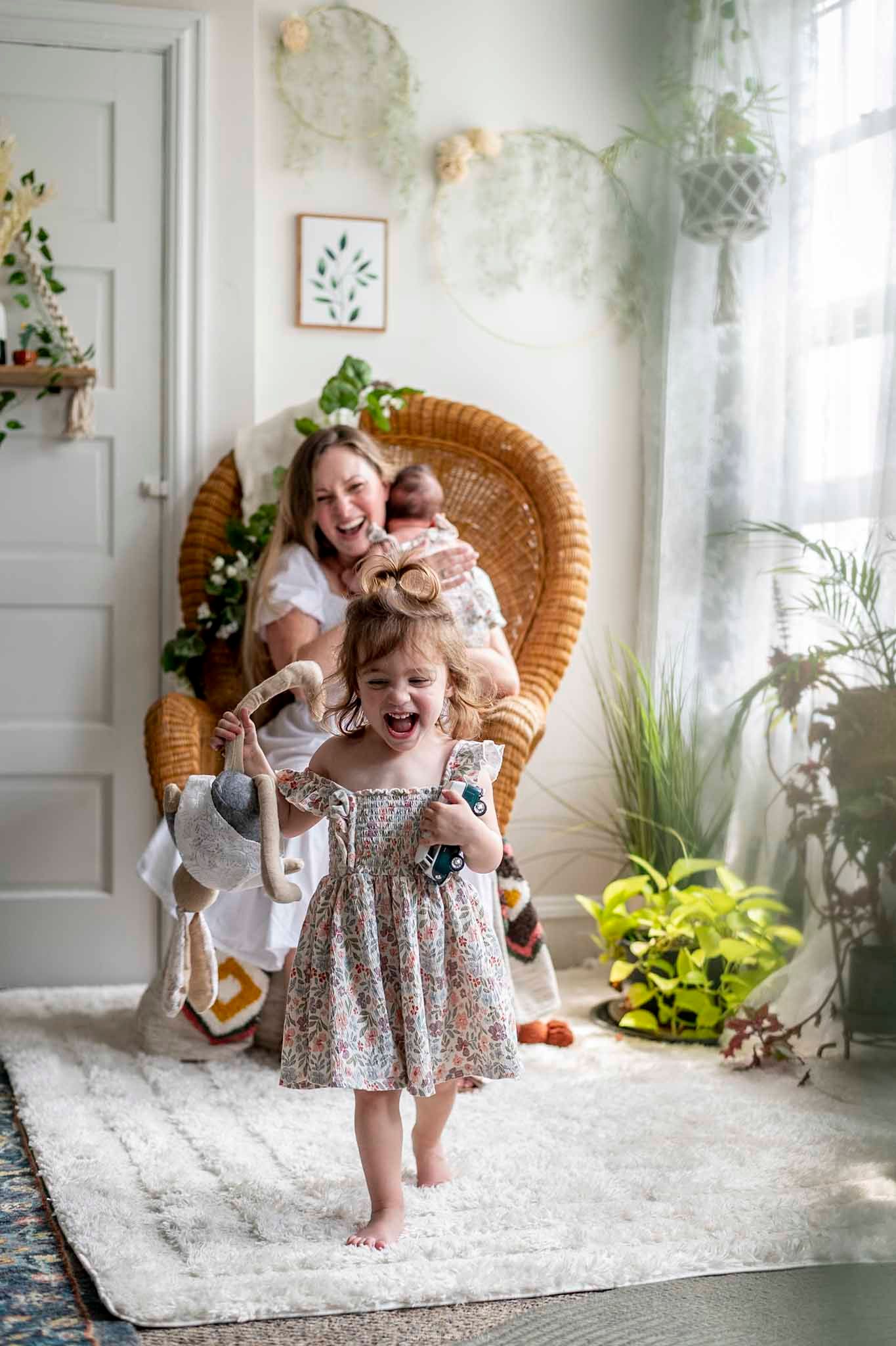 A joyful toddler girl in a floral dress laughs while a mother holds a baby in a cozy, plant-filled boho nursery.