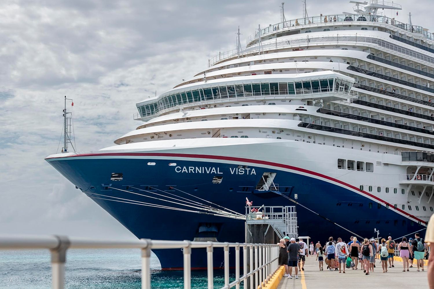 Carnival Vista cruise ship docked at a tropical port with passengers walking along the pier on a cloudy day.
