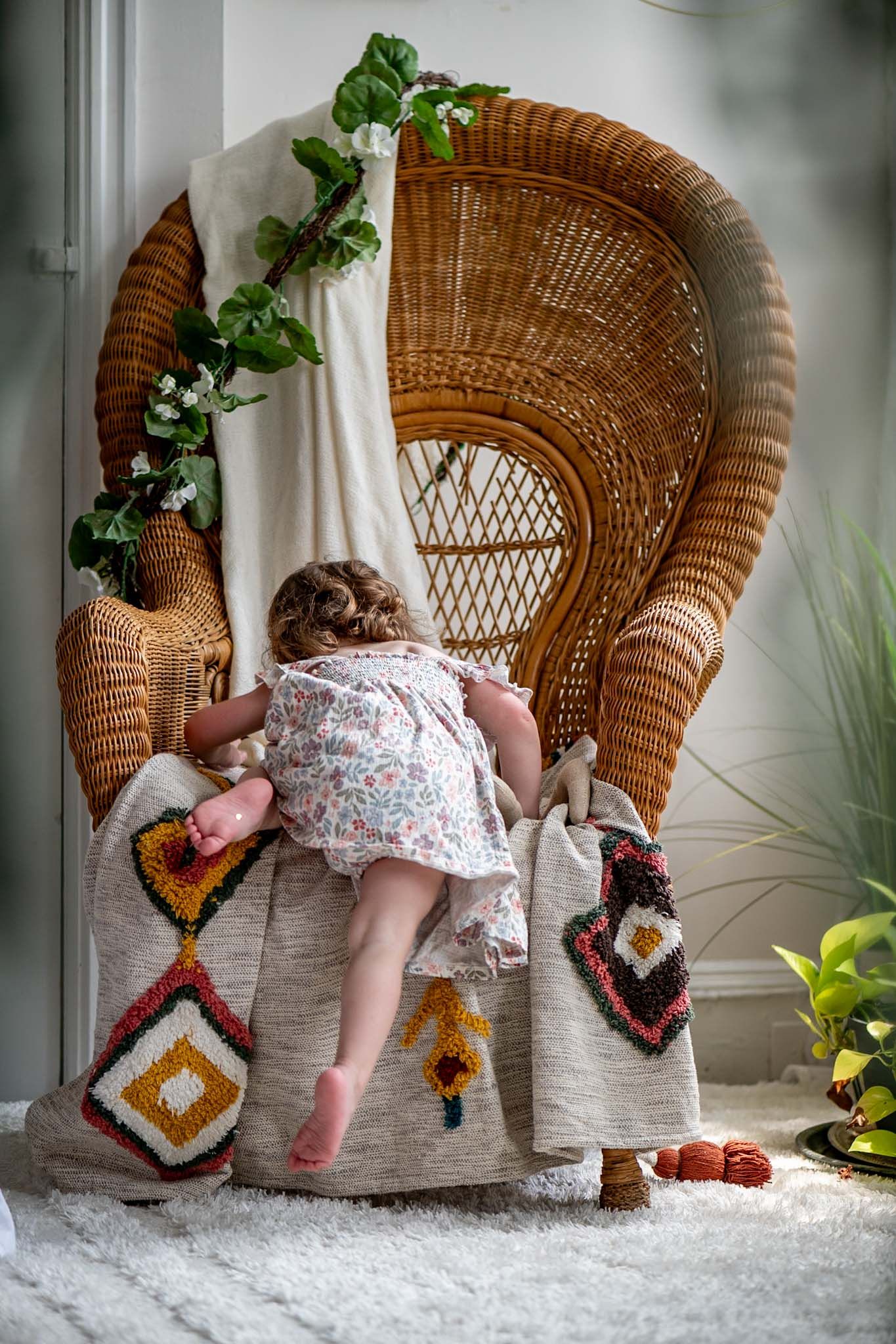 A wicker peacock chair sits against a white wall decorated with hanging plants and colorful textiles.