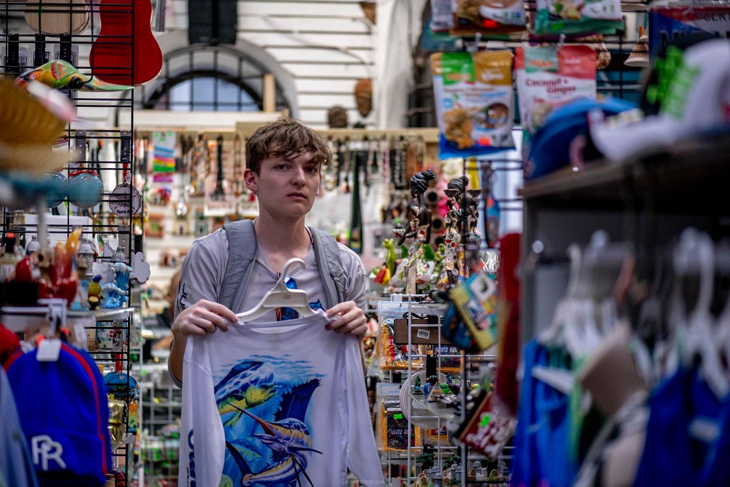 Young man browsing colorful souvenirs at an outdoor market stall, holding a graphic t-shirt.