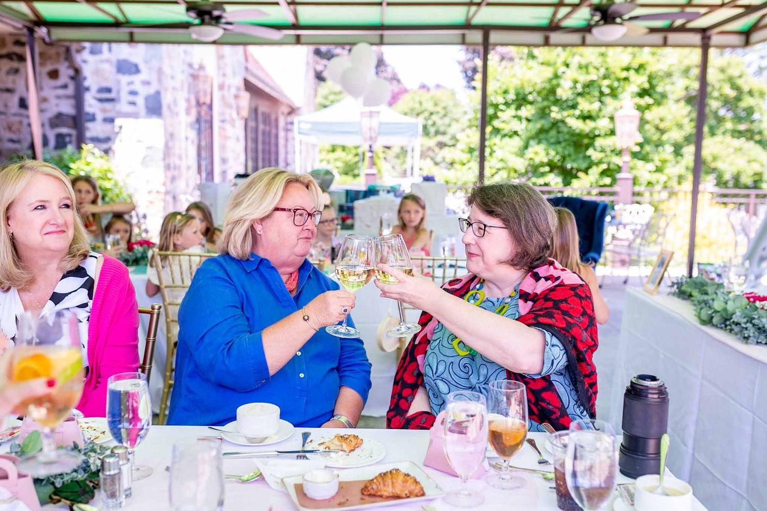 Two women toasting wine glasses at an outdoor brunch event with guests in background.