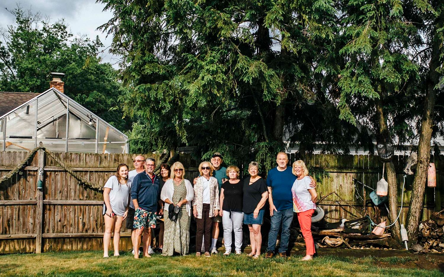 Large family group posing together in a backyard near a wooden fence and tall green trees on a cloudy day.