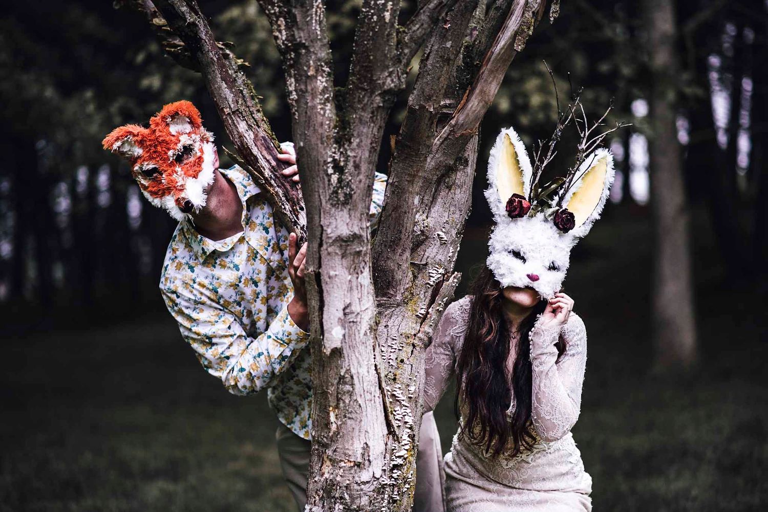 Two people wearing animal masks, a fox and a bunny, pose dramatically around a tree in a dark forest setting.