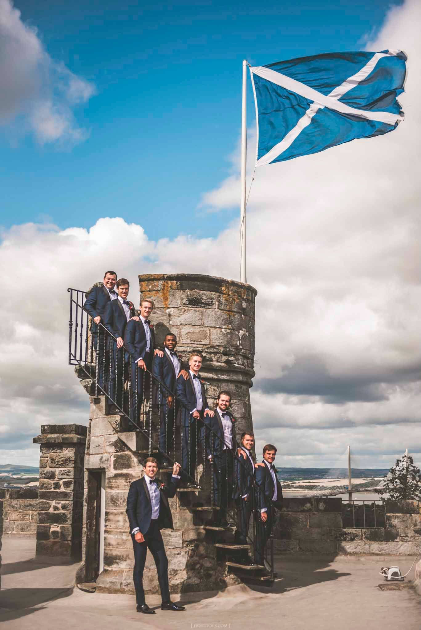 Groomsmen in navy suits pose on castle stairs with Scottish Saltire flag flying overhead under blue sky.