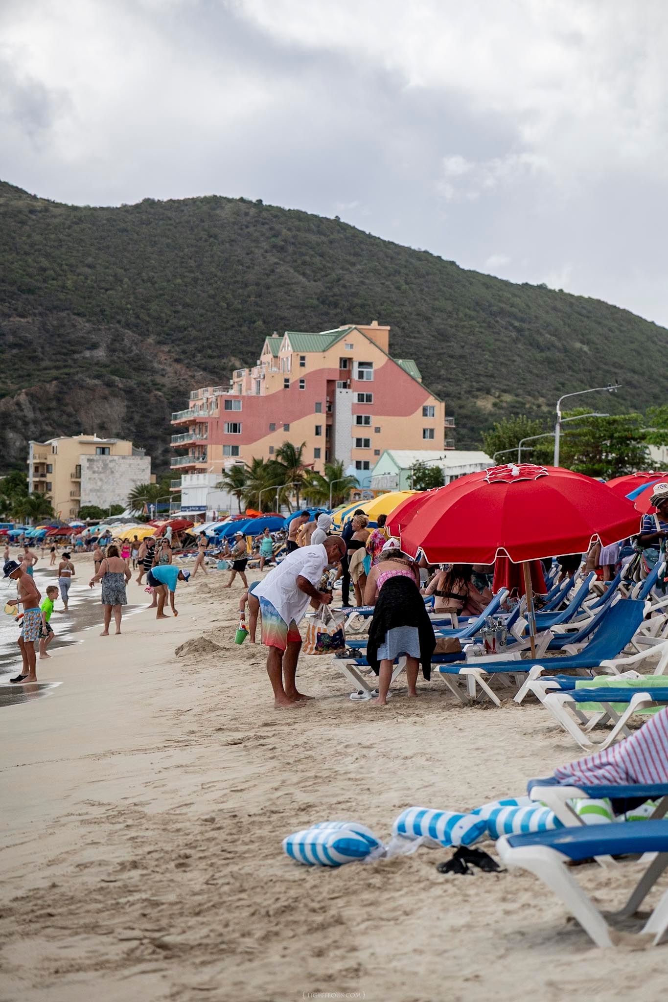 Crowded Caribbean beach with colorful umbrellas, lounge chairs, and tourists against a lush green hillside backdrop.
