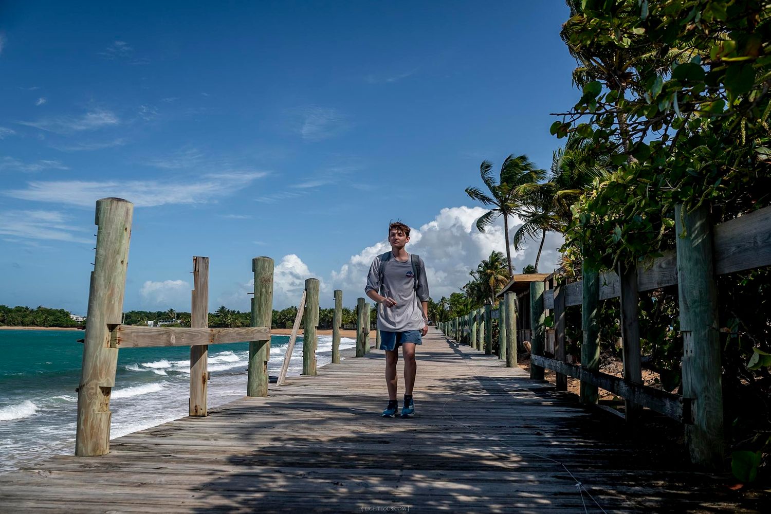Man walking on a wooden boardwalk beside a tropical beach with palm trees and blue sky.